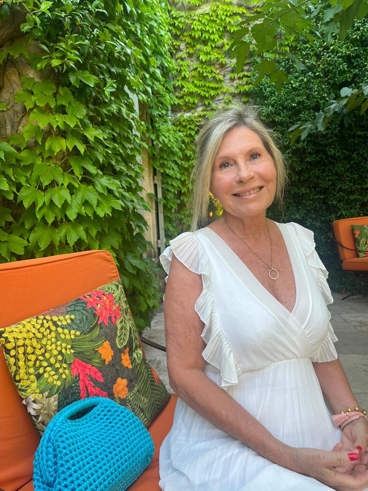 A smiling middle-aged woman with short blonde hair, wearing a green blouse with ruffled edges and a silver necklace, posing outdoors with green foliage in the background.