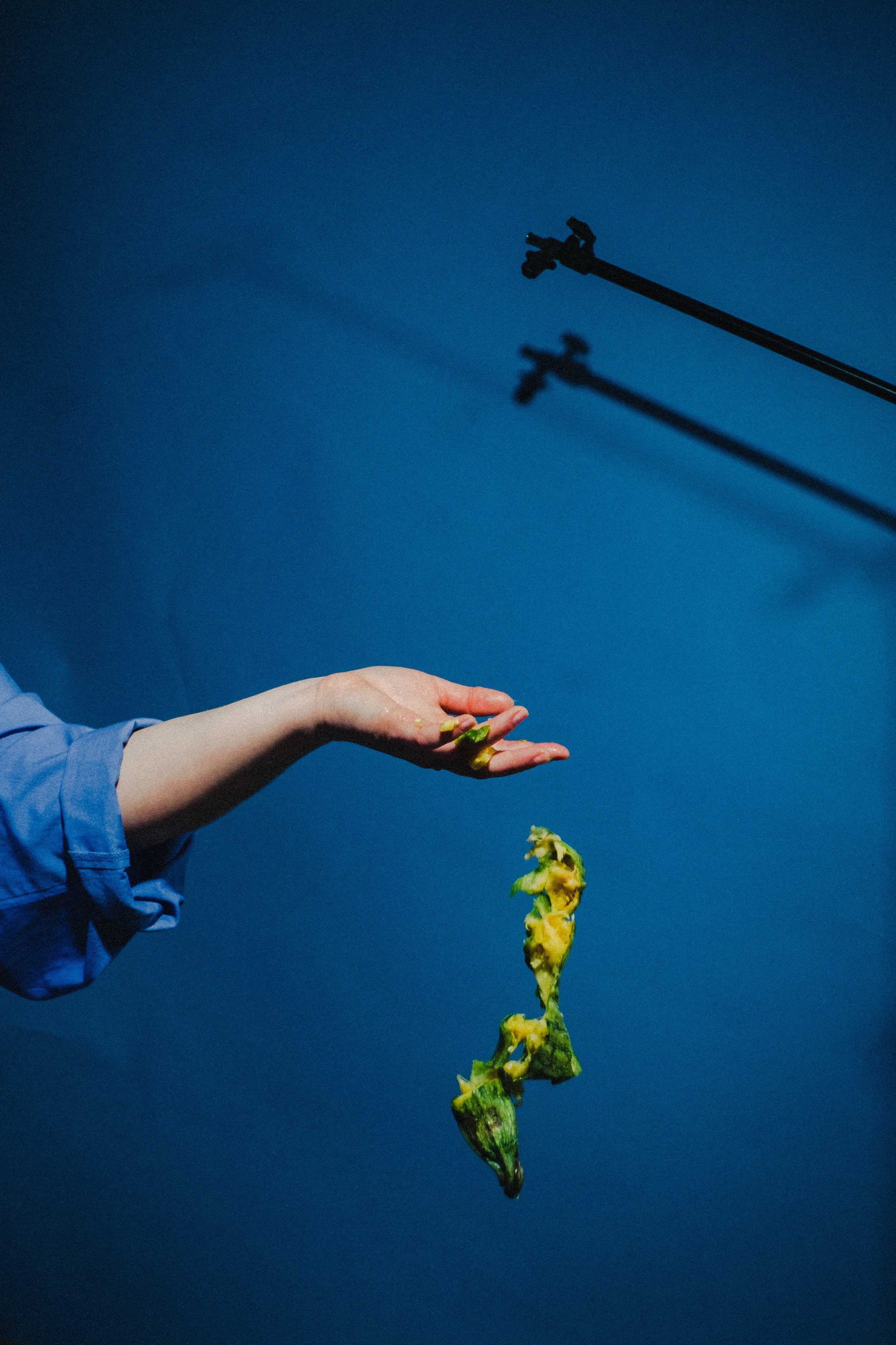 A person in a blue shirt is tossing a green and yellow object, possibly food, against a blue background with two overhead stage lights casting shadows on the wall.