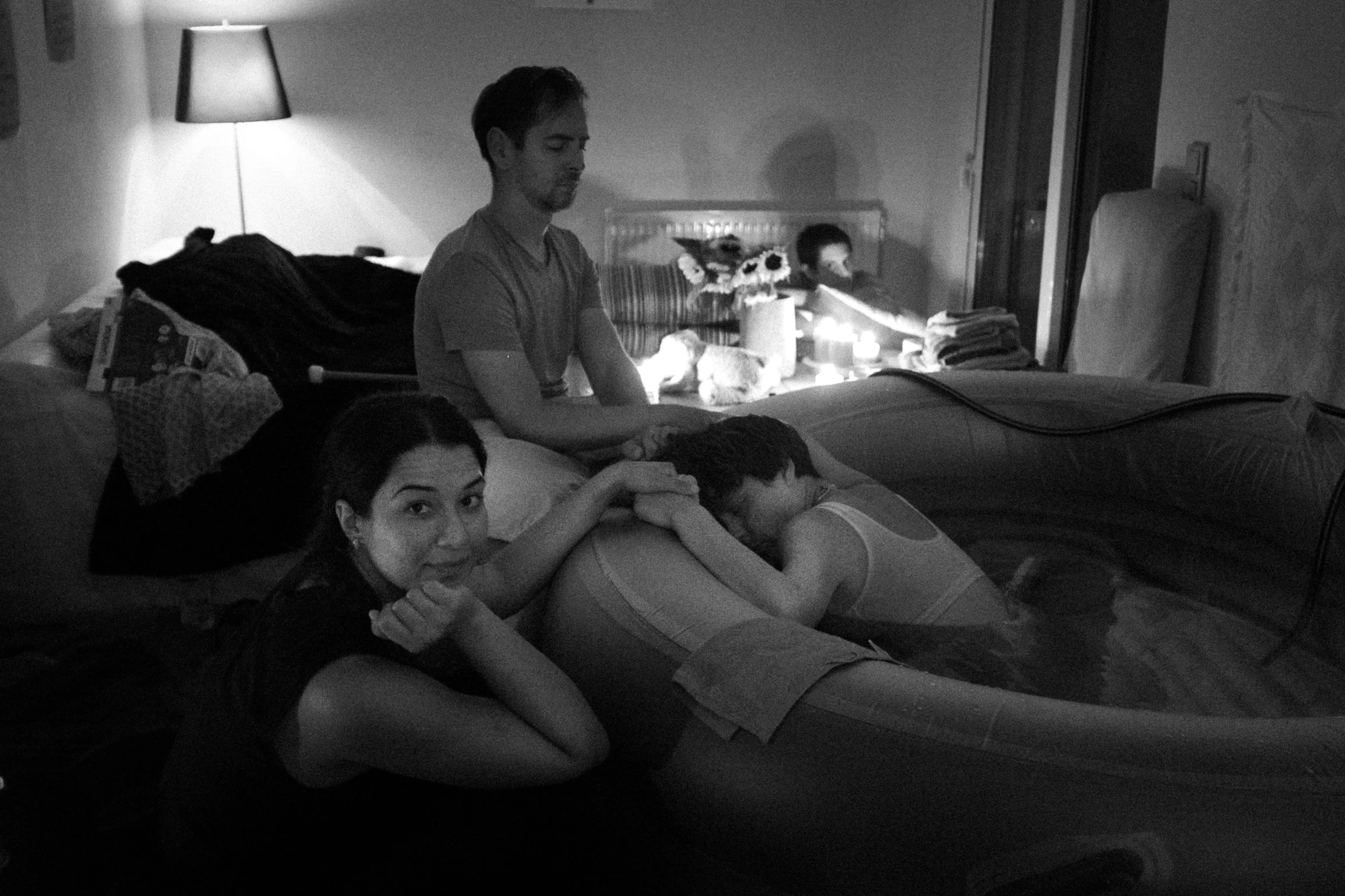 A group of people, including a woman and children, in a dimly lit room with candles and flowers, some resting on a bed and others sitting nearby.