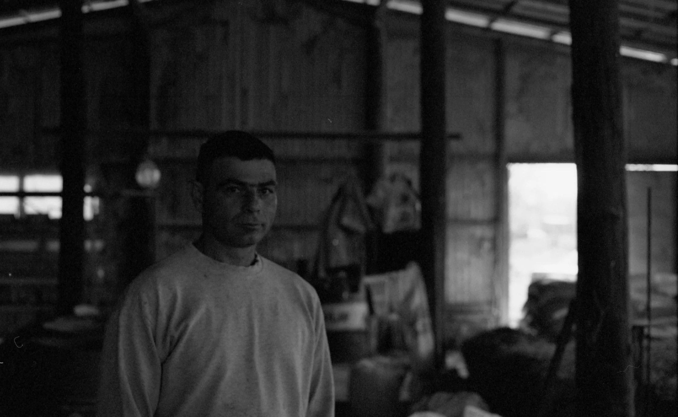 A black and white photo of a man standing inside a rustic wooden structure, looking at the camera.