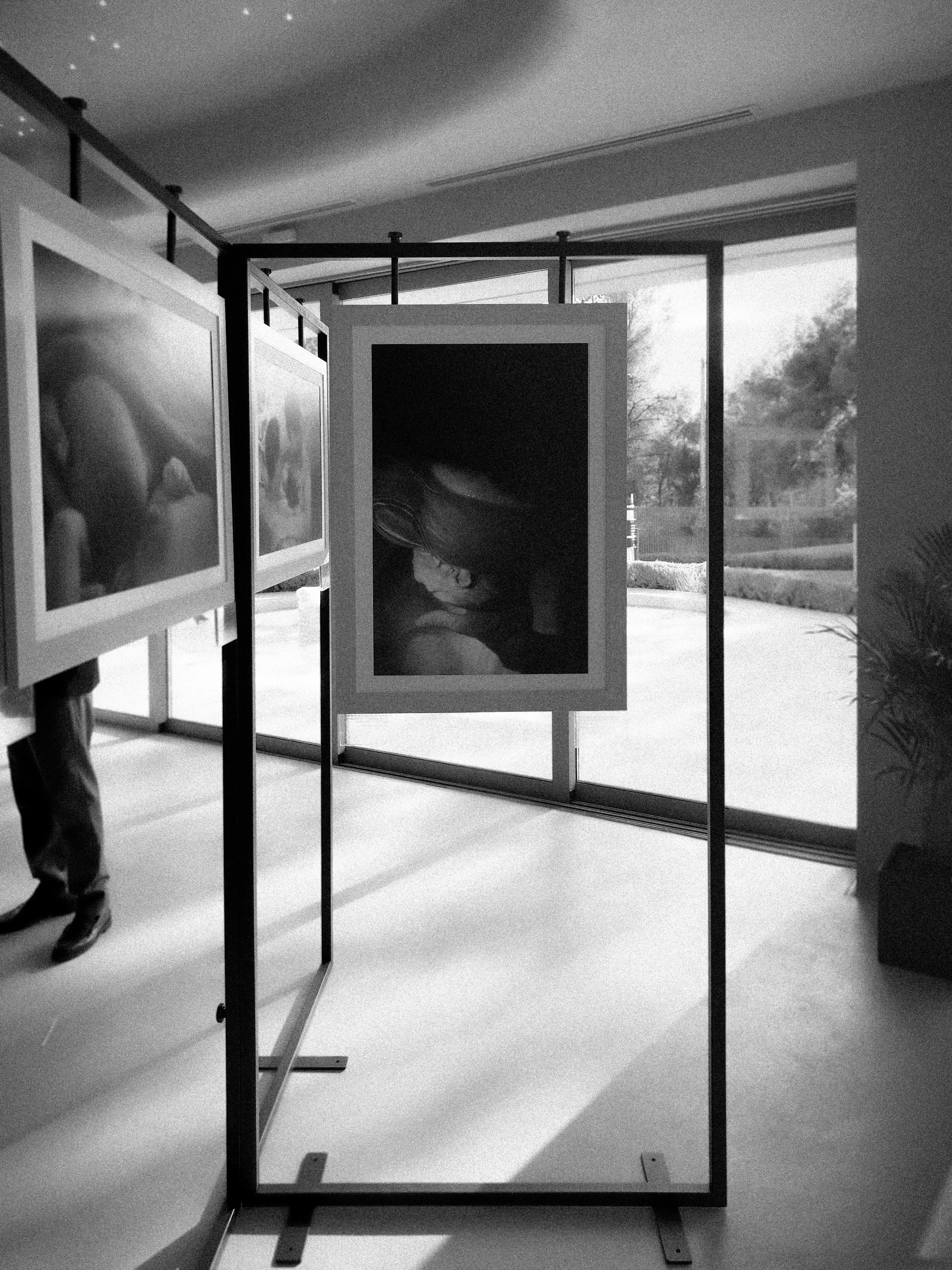 Black and white photo of an art gallery with framed photographs on display, glass windows in the background, and a person partially visible on the left.