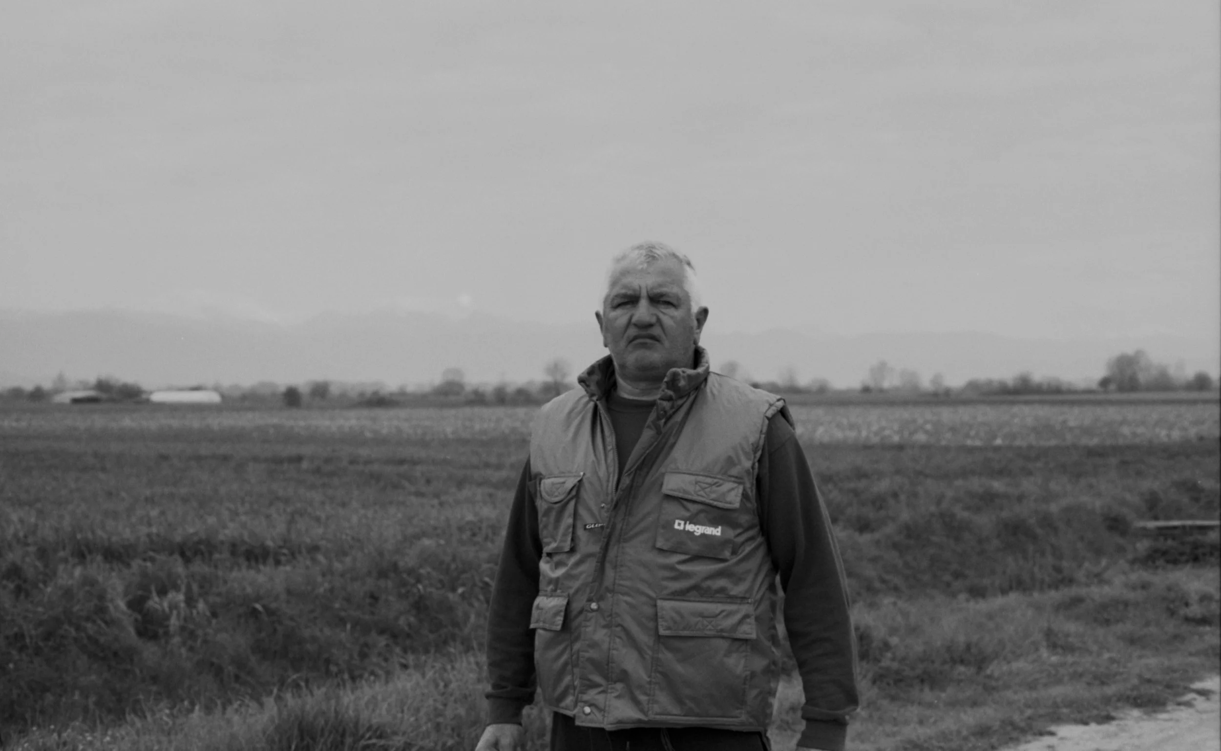 A man with gray hair wearing a vest standing outdoors in a field with mountains in the background, black and white photograph.