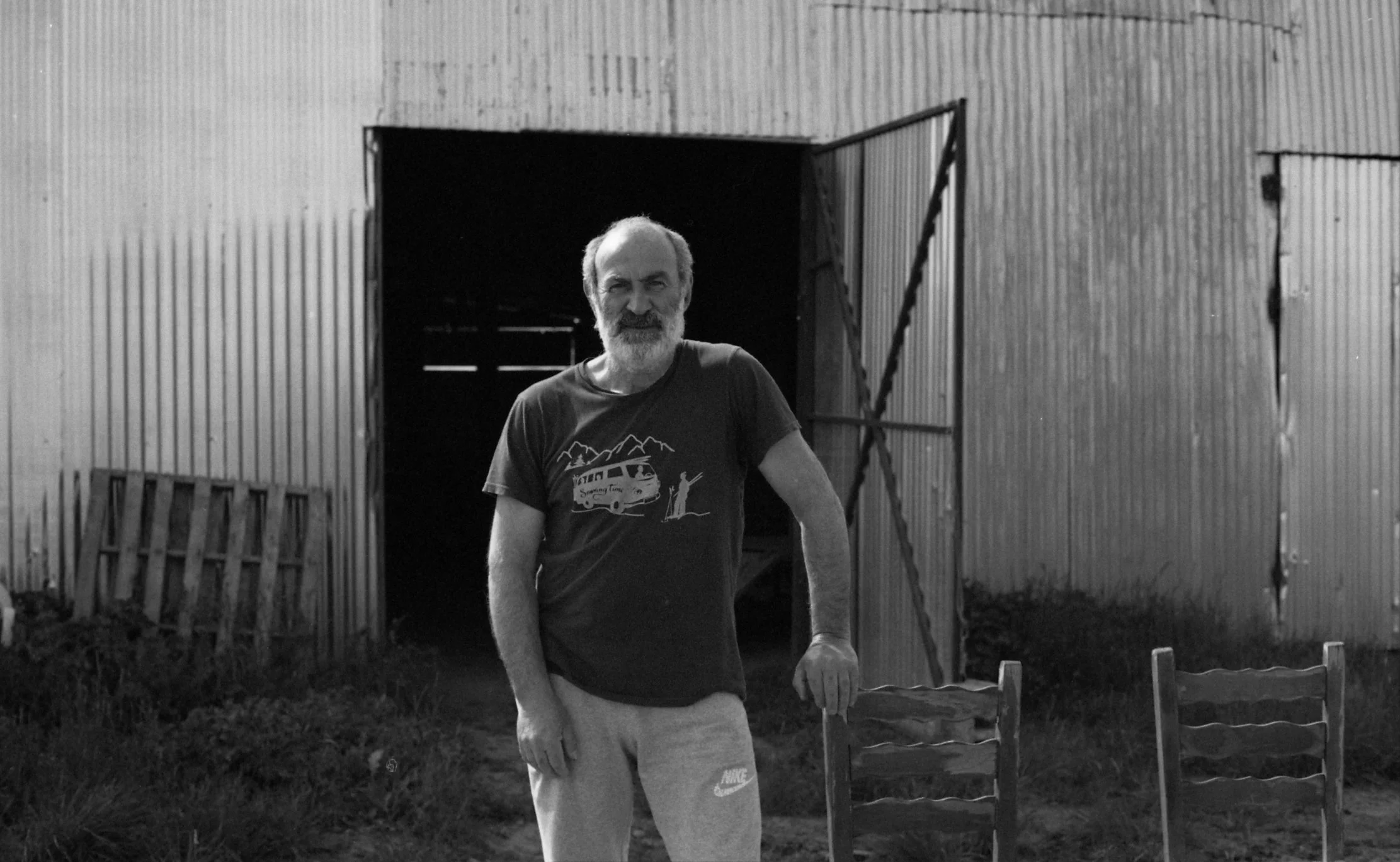 A man with gray hair and a beard standing outdoors in front of a corrugated metal building, leaning on a wooden chair, smiling. There are additional chairs and a ladder visible.
