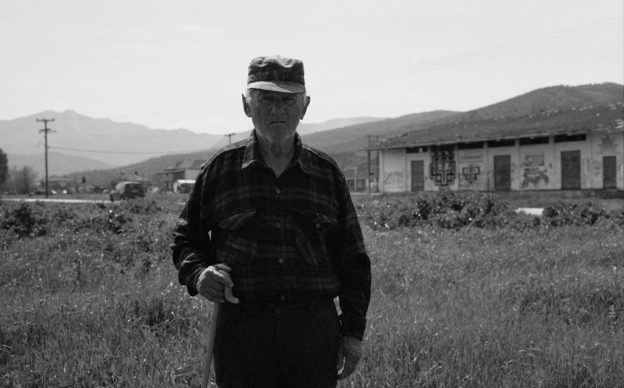 A black and white photo of an elderly man standing outdoors in a grassy area with hills and mountains in the background, wearing a cap and plaid shirt.