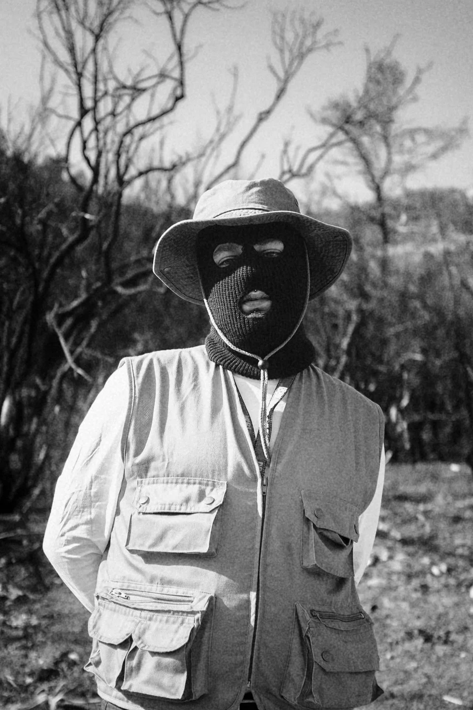 Person wearing a wide-brimmed hat, ski mask, and layered vest outdoors in a wooded area.