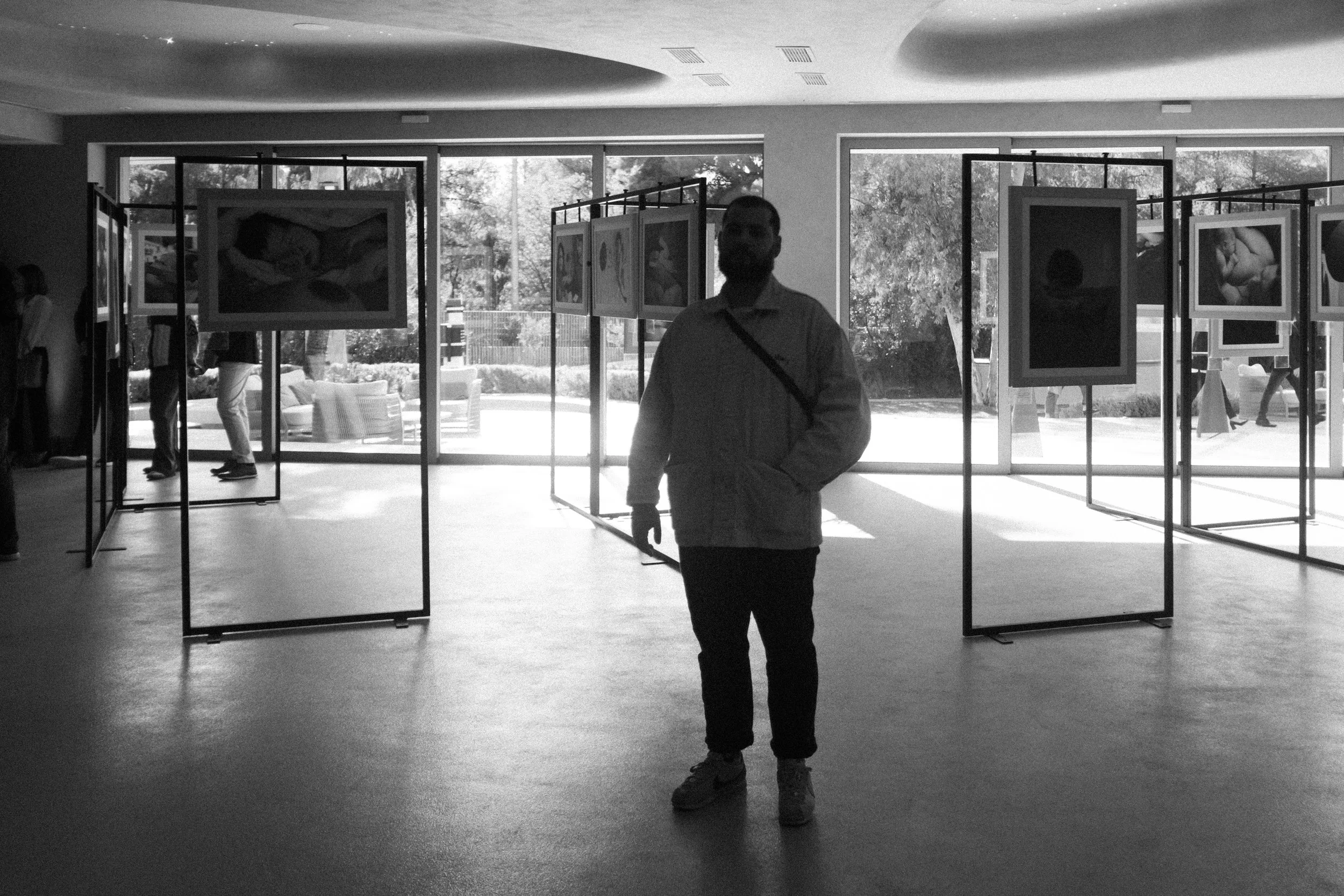 A person standing inside an art gallery with artwork displayed on partition walls, with large windows in the background showing trees and outdoor seating.