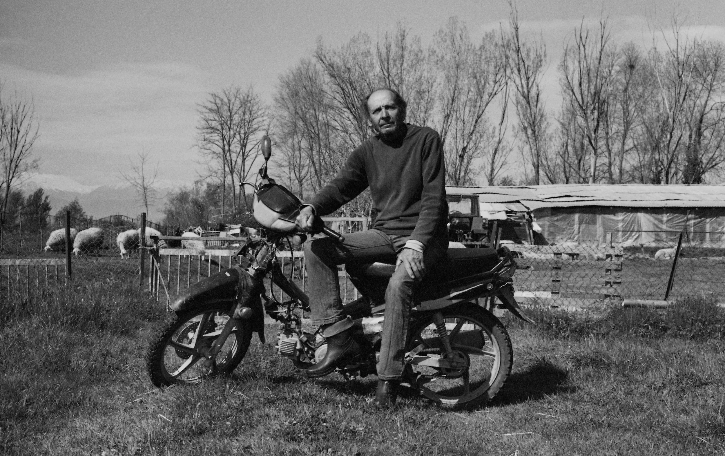 A man sitting on a motorbike in a grassy area with trees and a fence in the background, black and white photo.