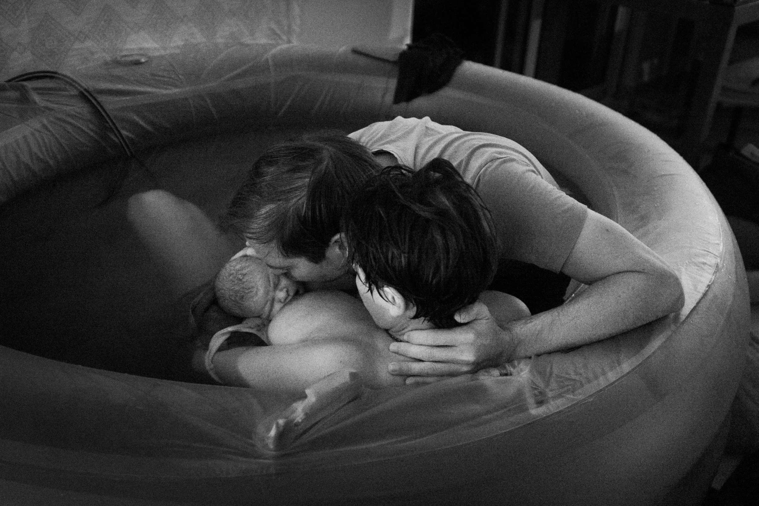 Two adults holding a newborn baby in an inflatable pool, sharing a tender moment in black and white.