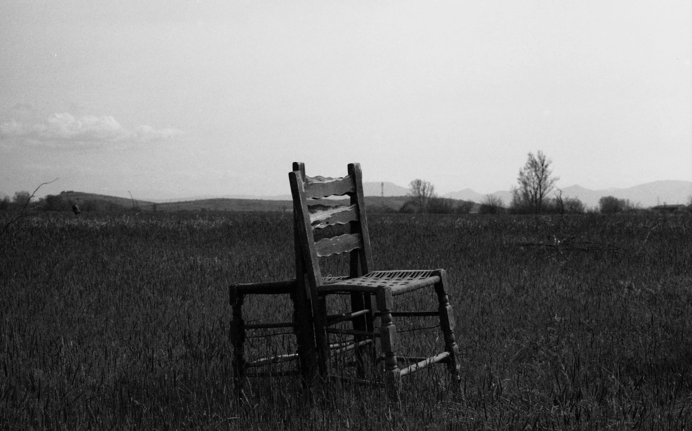 An old wooden chair in an open field with hills and trees in the background, black and white photograph.