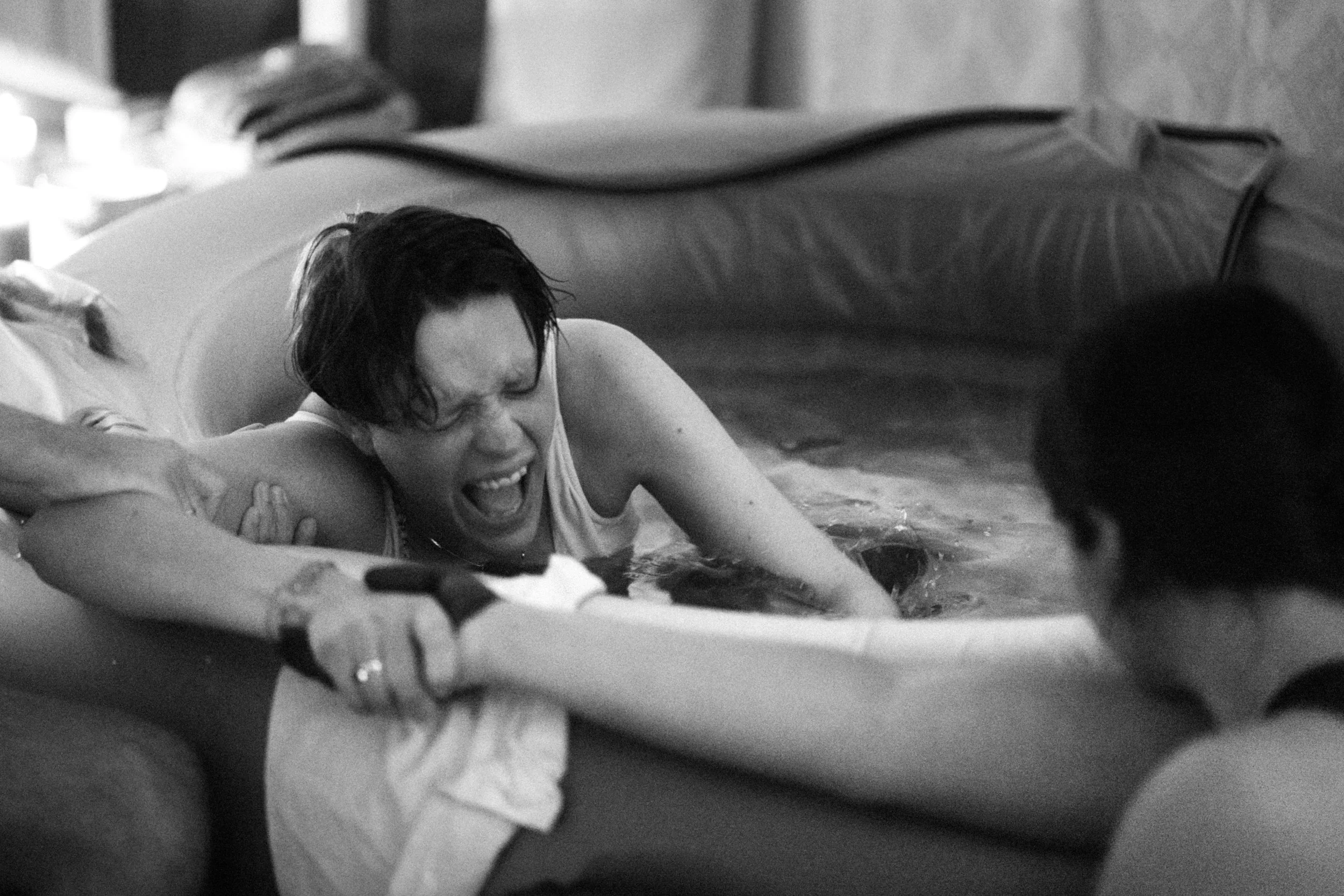 A woman struggling in a pool, grimacing and holding onto the side as she is assisted by a person outside the pool.