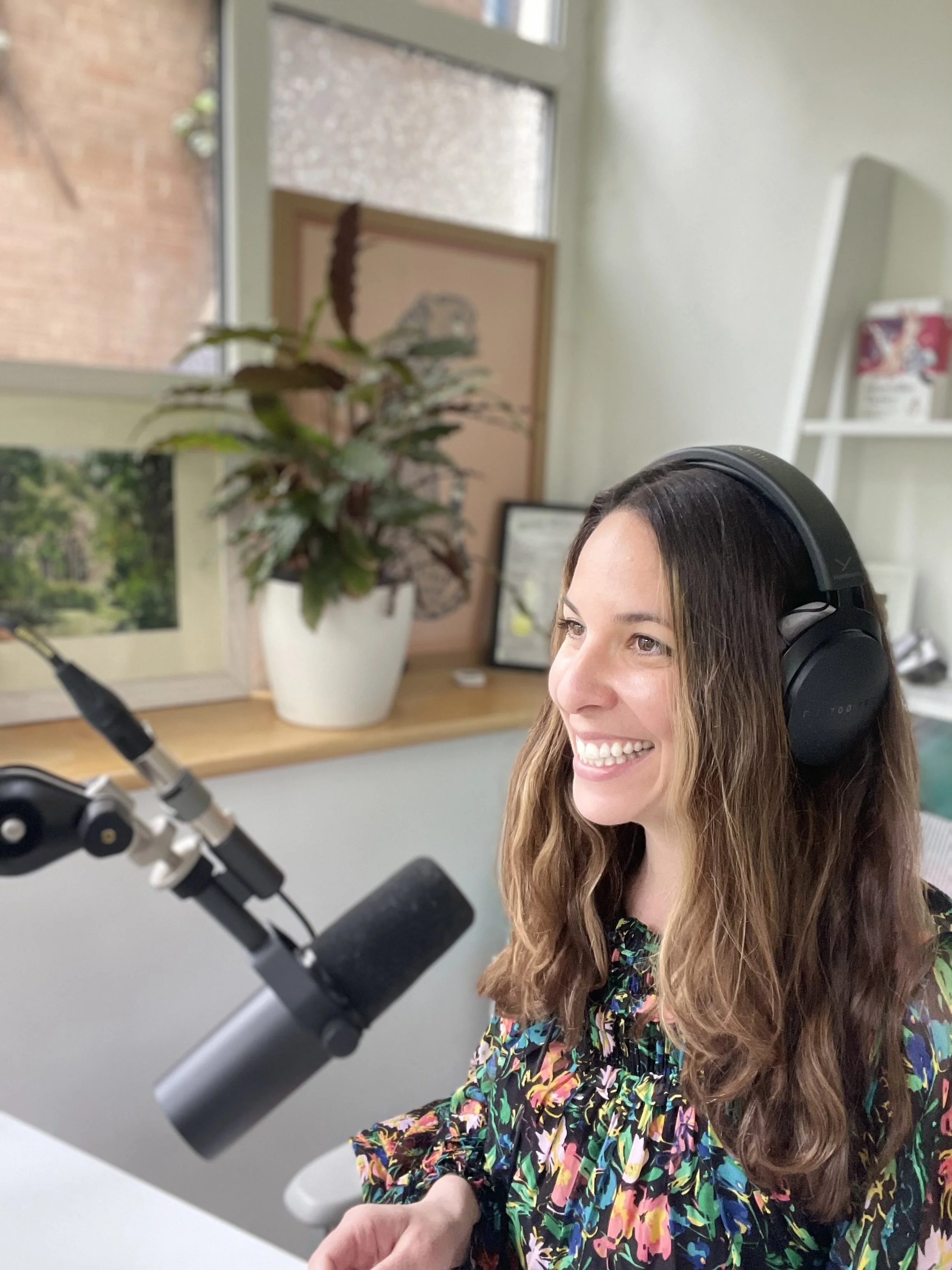 A woman with long wavy hair, wearing headphones, smiling while speaking into a microphone in a home office or recording studio.
