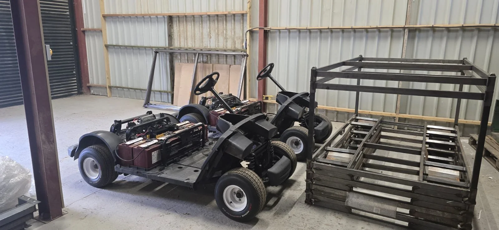 Two electric golf carts and a large metal cart frame are inside a warehouse with metal siding walls.
