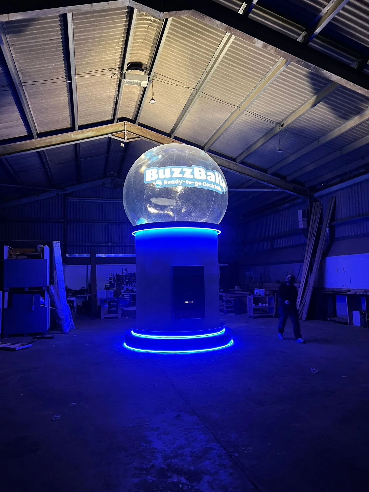 A large illuminated globe-shaped display with the words 'BuzzBall Ready-to-go Cocktails' inside a warehouse or industrial space, with blue lighting, metal roof, and a person standing near the right side.