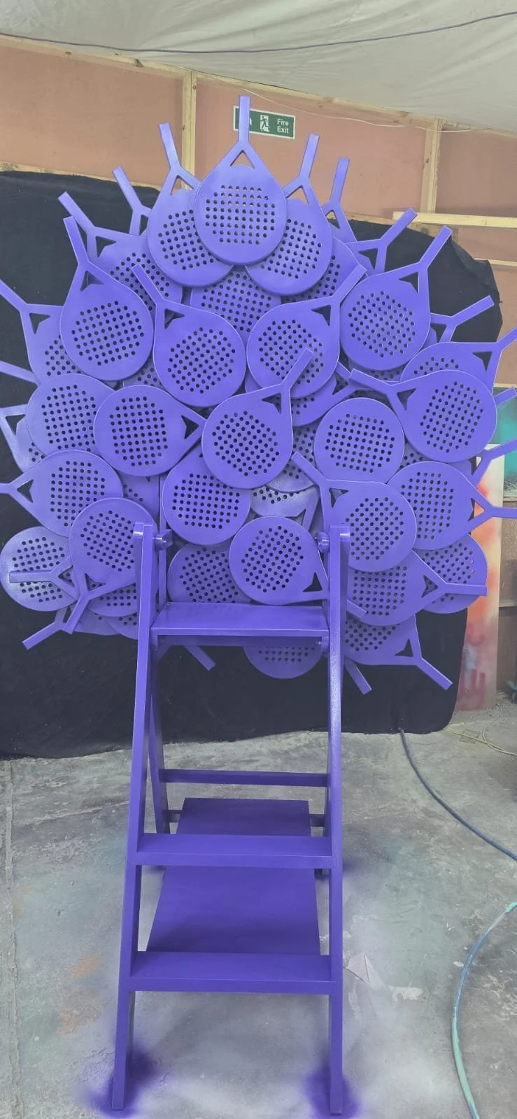 Purple wooden chair in front of a wall of purple watering cans arranged to resemble a peacock.