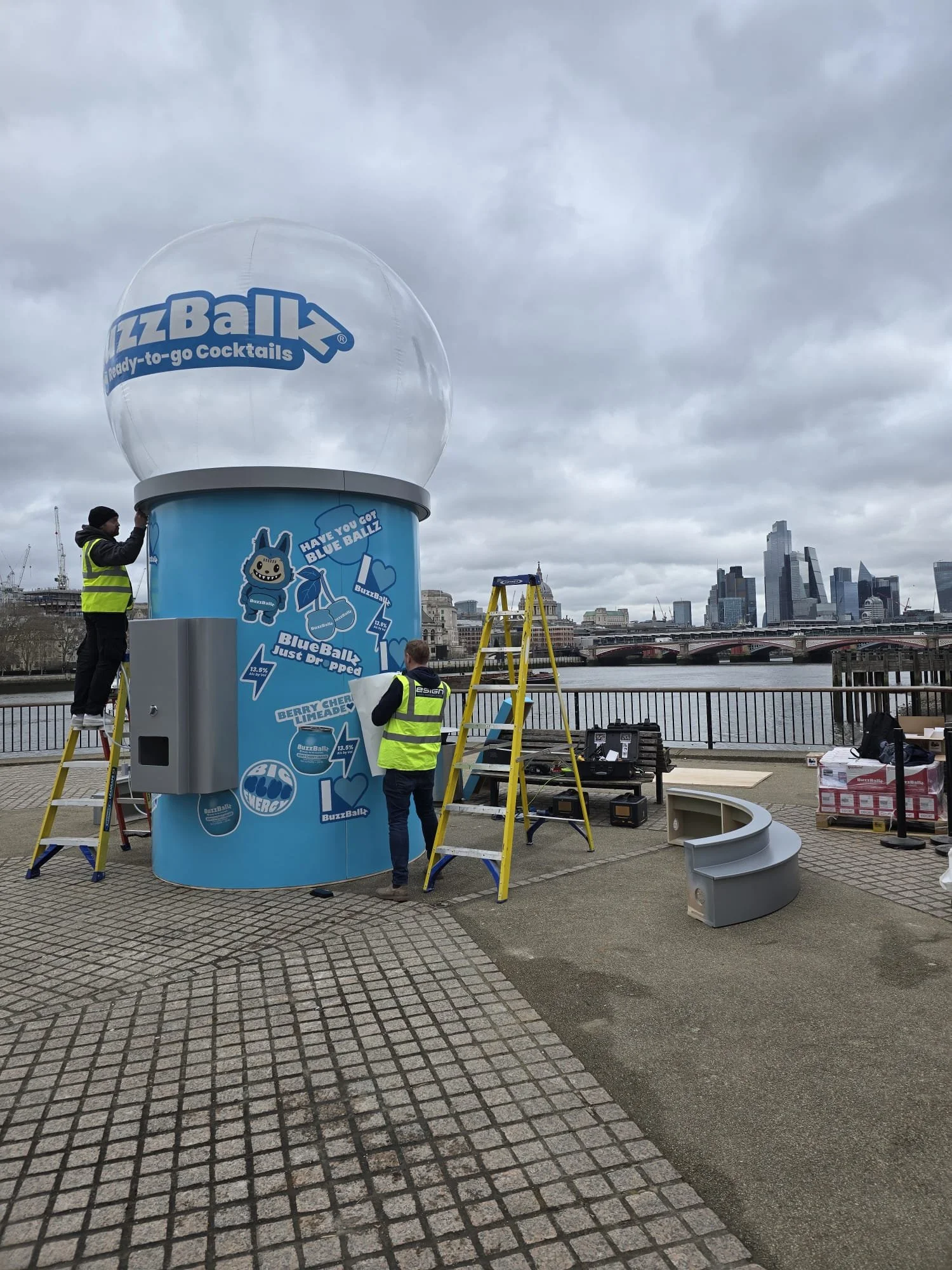 Installation of a BuzzBall promotional display on a riverside promenade, with workers setting up a large blue cylindrical sign featuring BuzzBall branding and stickers, under a cloudy sky with city skyline in the background.