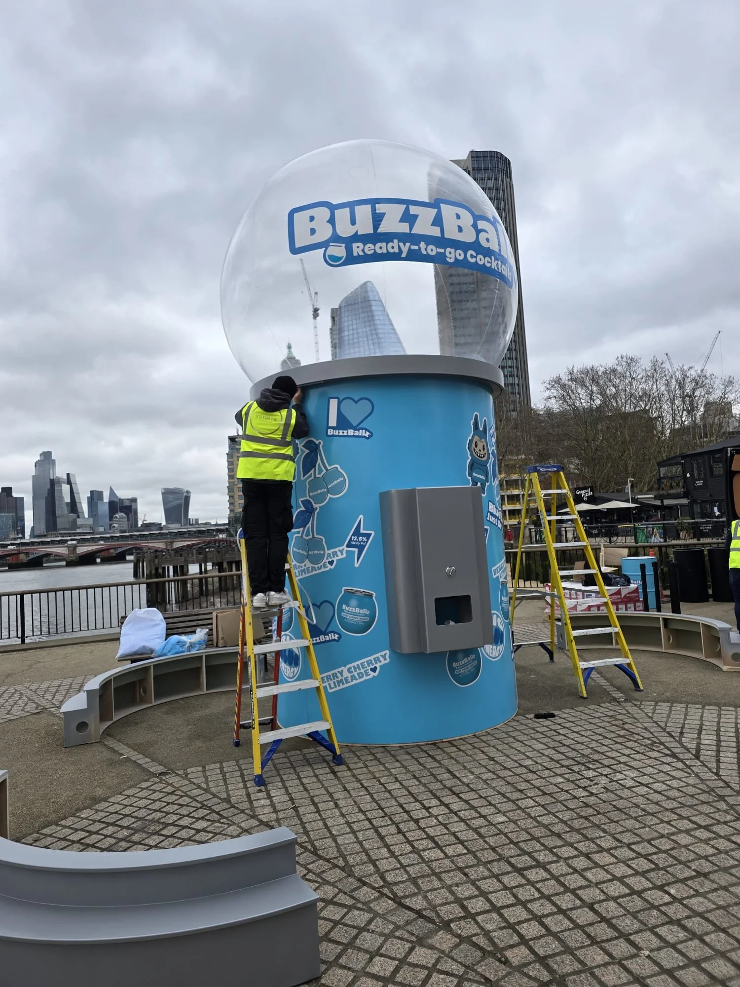 Workers installing a BuzzBallz promotional display on a city riverside promenade. The display features a large, transparent bubble with the BuzzBallz logo and the phrase 'Ready-to-go Cocktails'. The background shows tall buildings and overcast skies.
