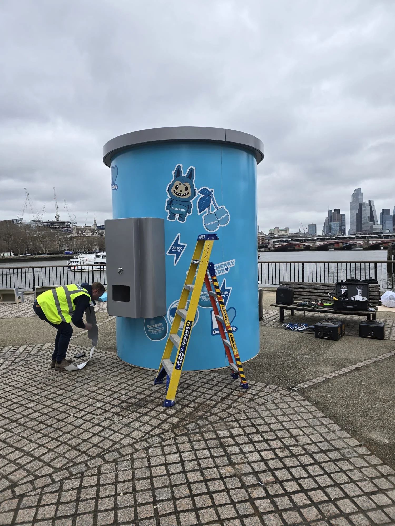 A large blue cylindrical promotional booth with cartoon monster and flavor logos, a person in a yellow safety vest setting up equipment nearby, and a city skyline with river in the background.