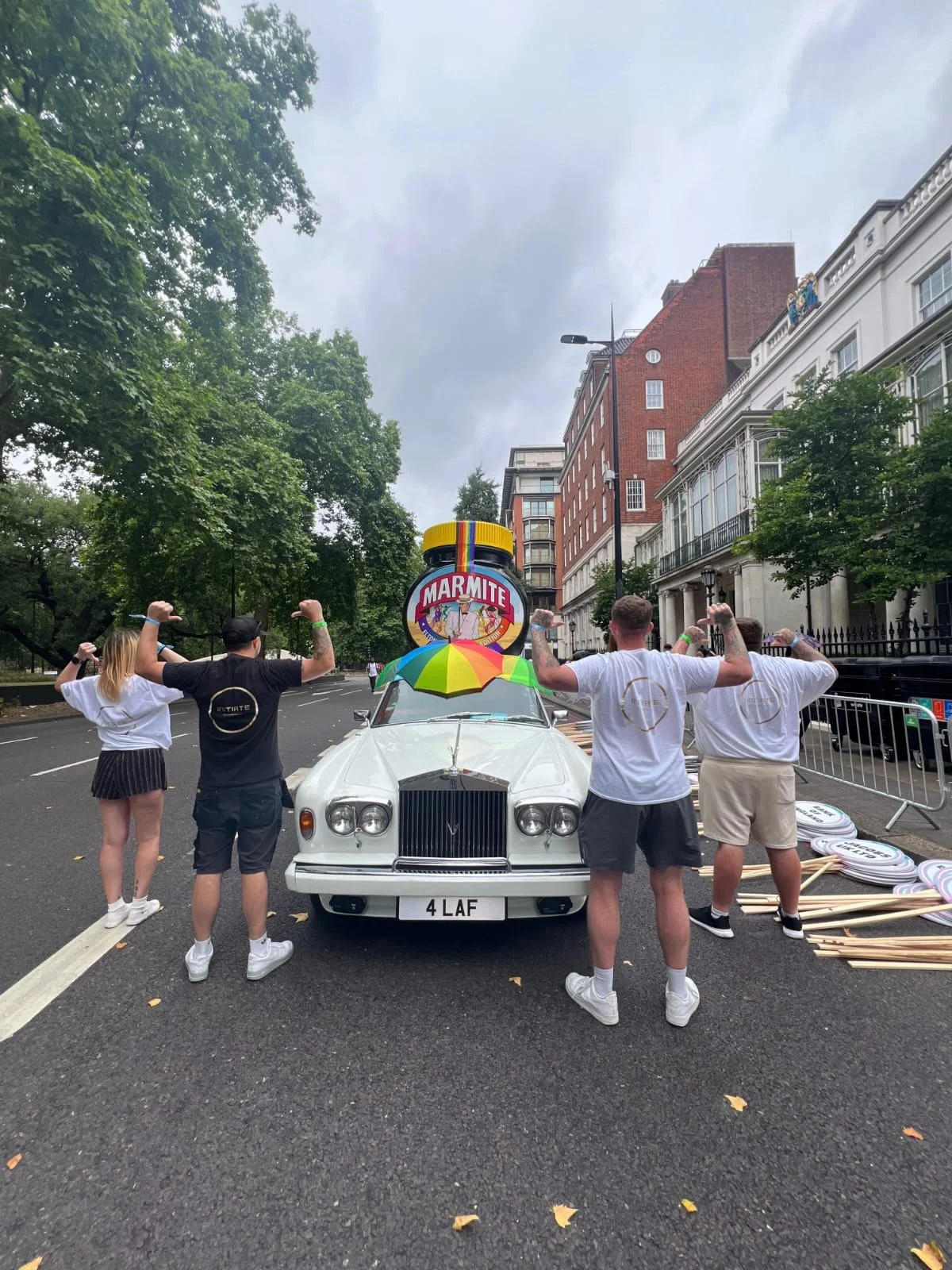 Four people standing in front of a white vintage car with a Marmite sign on top of the car, holding their arms up in a flexing pose, on a city street with buildings and trees in the background.