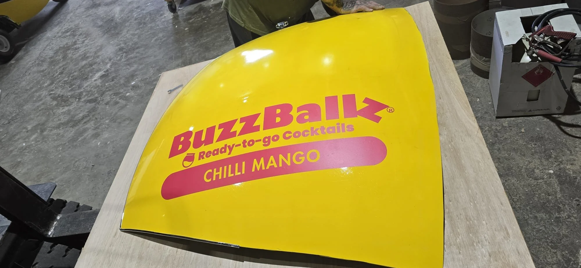 Yellow BuzzBall drink container with pink and red text, indicating it is a ready-to-go cocktail called Chilli Mango, placed on a light-colored wooden surface with some tools and a cardboard box nearby.