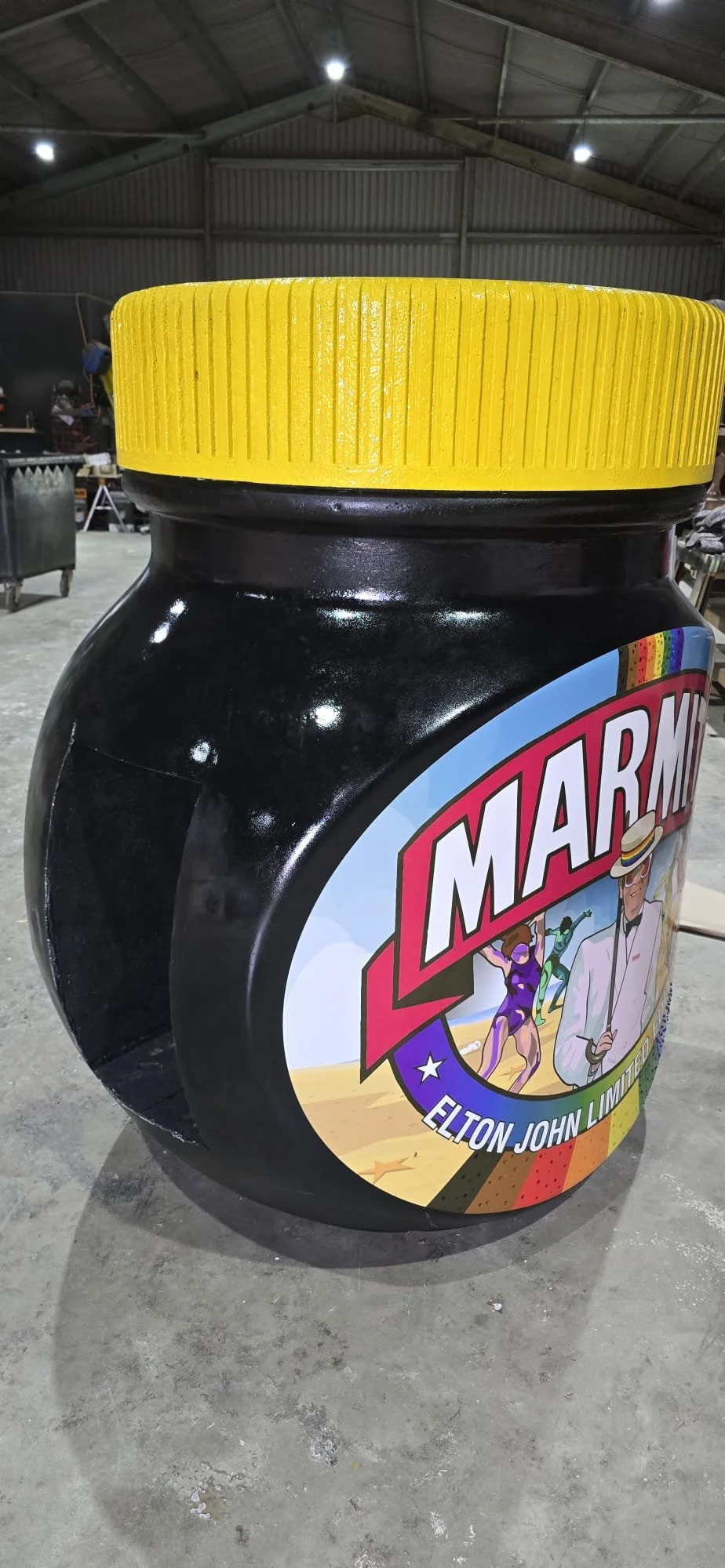 A large, black, plastic container shaped like a jar with a bright yellow lid, featuring a colorful label with the word 'Marmin' and illustrations of people at a beach.