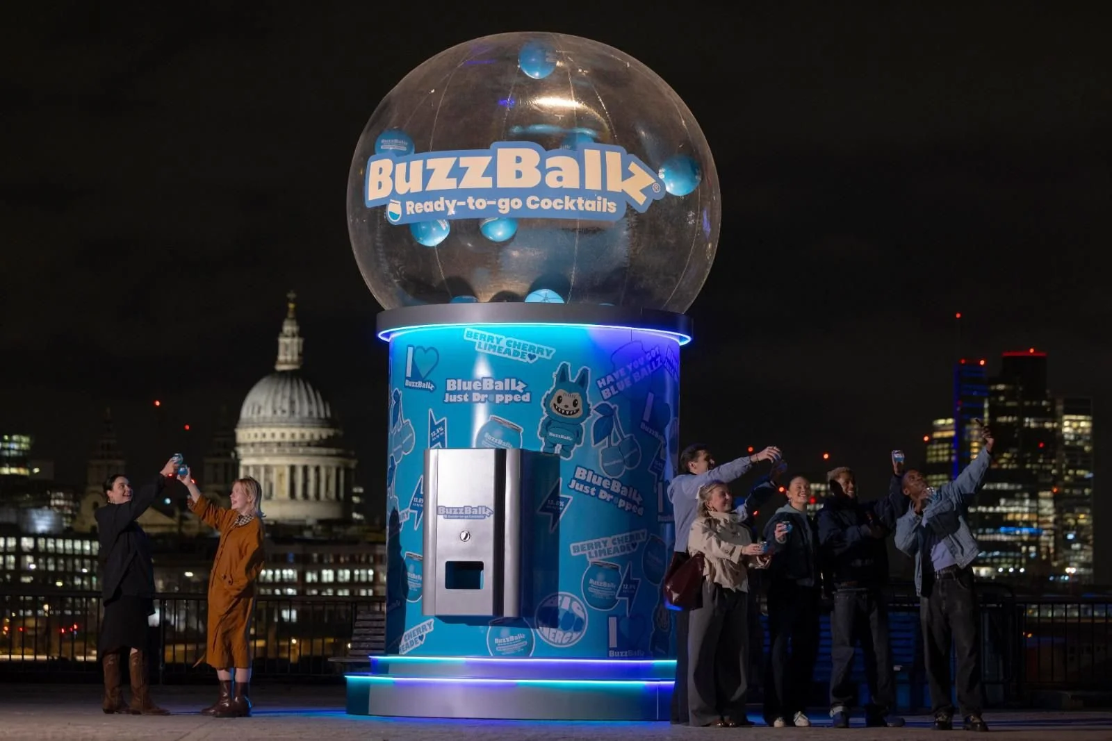 Night scene of a city skyline featuring a large BuzzBallk promotional display with a clear globe and blue themed branding. Several people are taking selfies and photos in front of the display, with St. Paul's Cathedral visible in the background.
