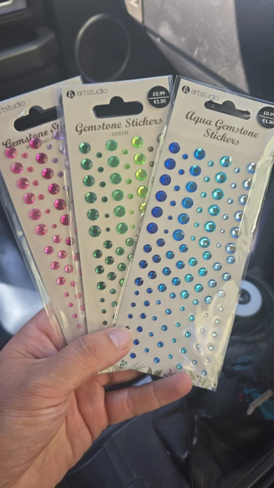 Hand holding three packs of Gemstone Stickers in pink, green, and blue colors inside a vehicle.