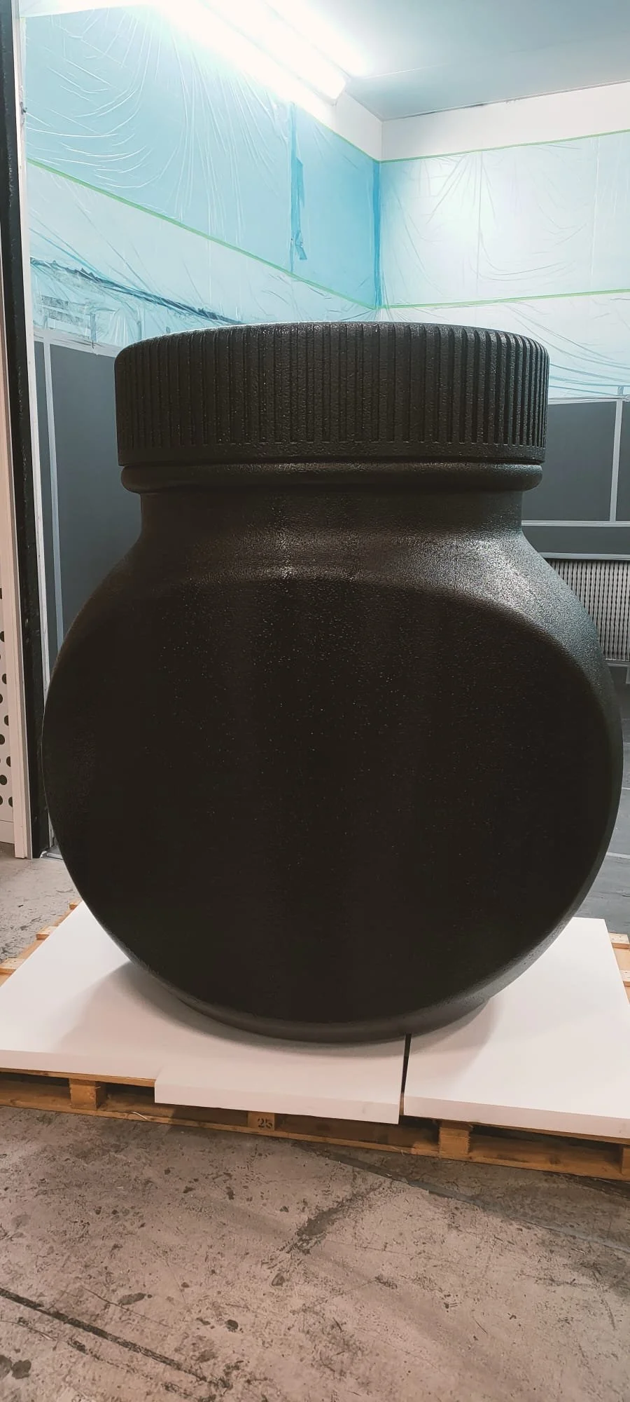 A large black object shaped like a rounded jar or vase with a textured screw-on lid, placed on a white platform and set against an industrial or warehouse background.
