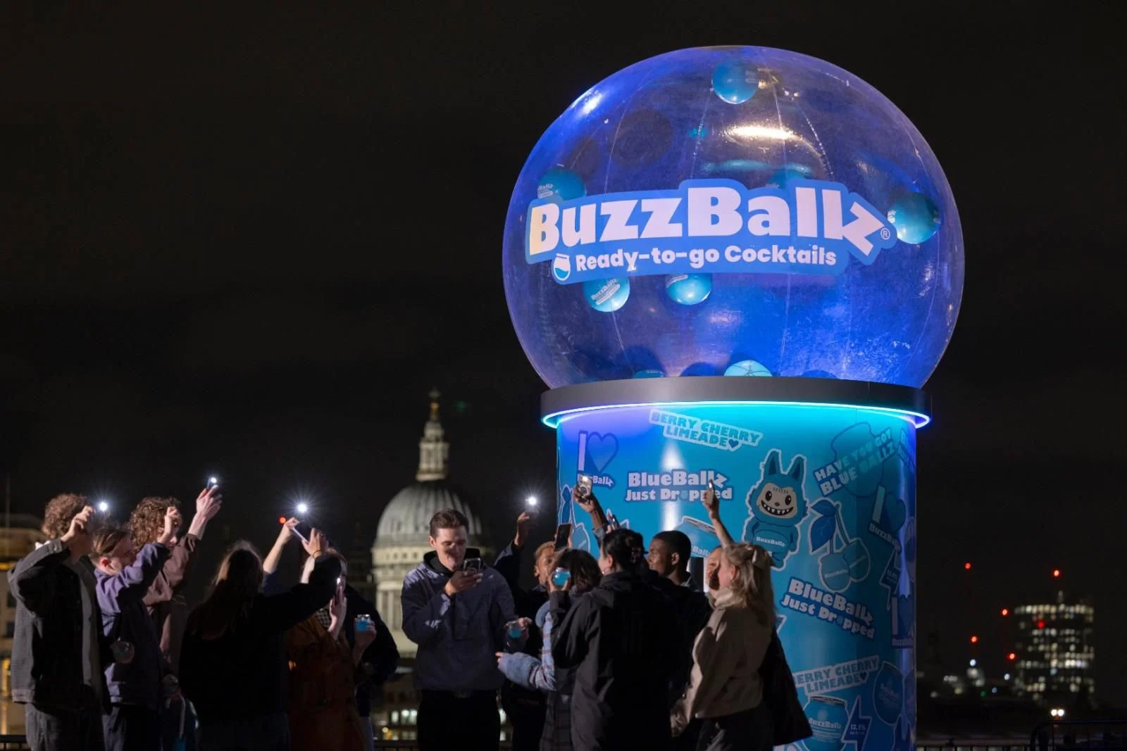 A group of people gathered around a large, illuminated BuzzBall display on a rooftop at night, with city buildings and a dome in the background.