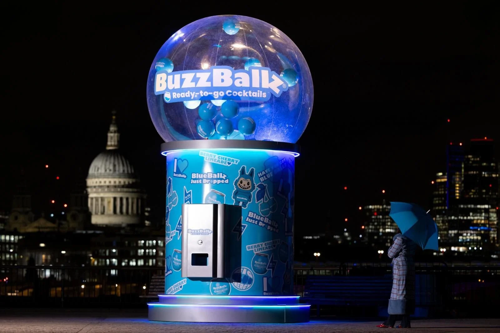 A brightly lit BuzzBall vending machine with a large transparent globe on top filled with blue balls, situated on a city rooftop at night. A person with an umbrella stands nearby, overlooking the city skyline and the dome of a historic building.