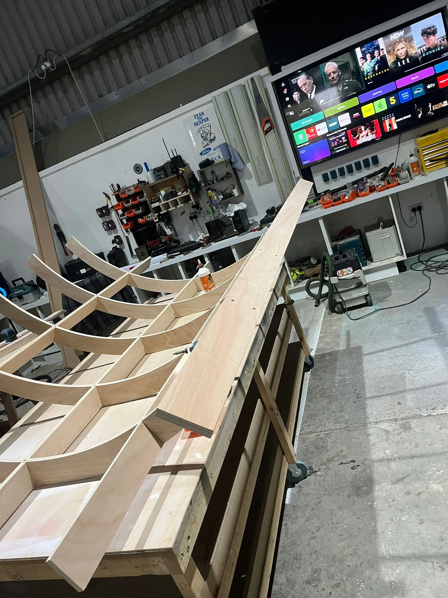Wooden frame structure  of THE BUZZBALLZ gatcha on a workbench in a workshop with tools and electronic equipment, and a large TV screen on the wall.