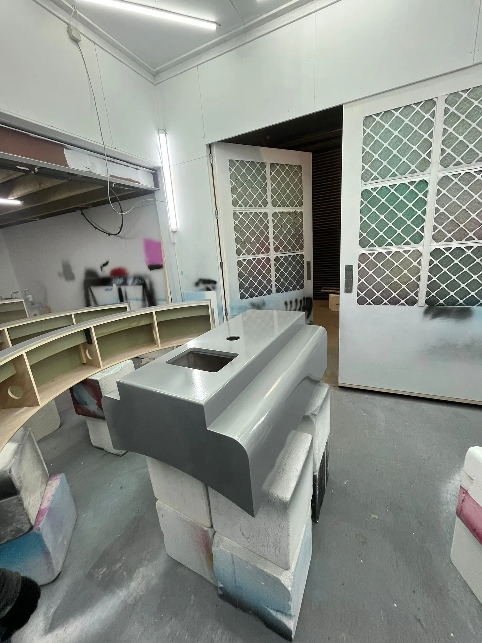 Industrial workspace with a gray painted piece of furniture on white packing Styrofoam blocks, a curved wooden piece with multiple compartments, and a sliding door with a diamond pattern.