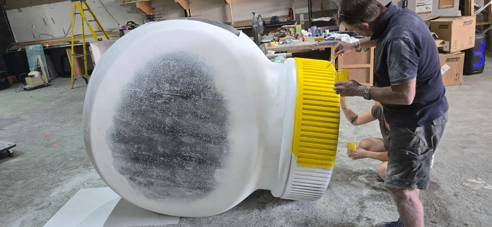Person painting a large, rounded, white sculpture or model with yellow details in a workshop. marmite