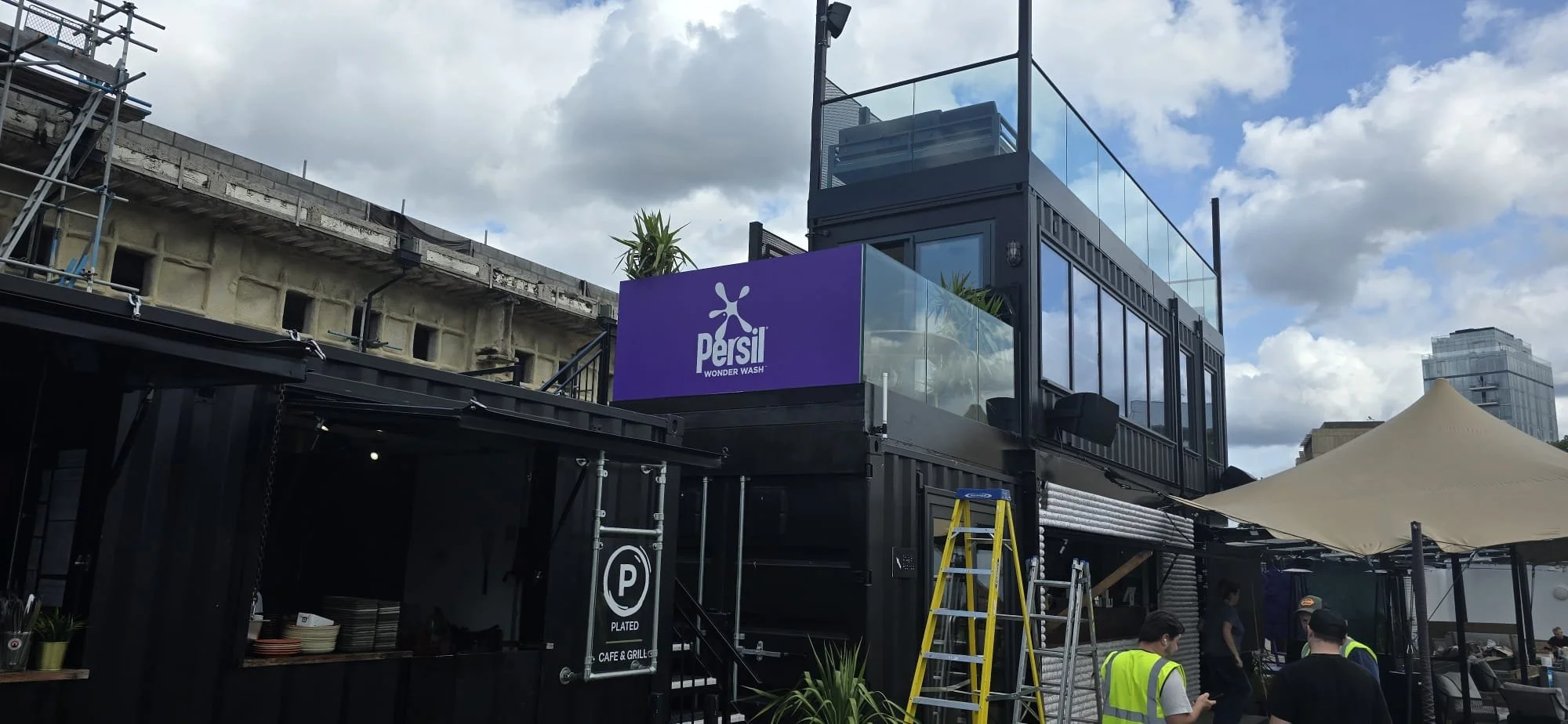 A two-story black building with glass windows, a large purple sign for Persil Wonder Wash, outdoor seating, a yellow ladder, and people wearing safety vests, set against a partly cloudy sky.
