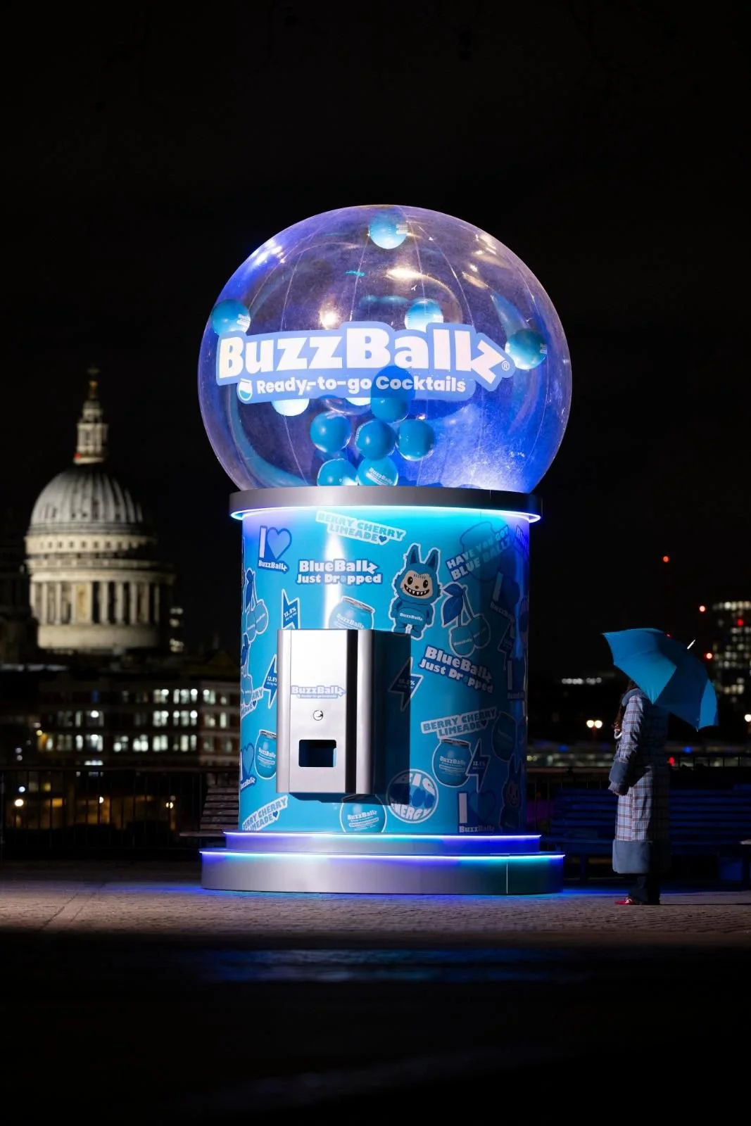 A BuzzBallz candy vending machine with a glowing blue design in an outdoor urban area at night, featuring a man with an umbrella standing nearby.