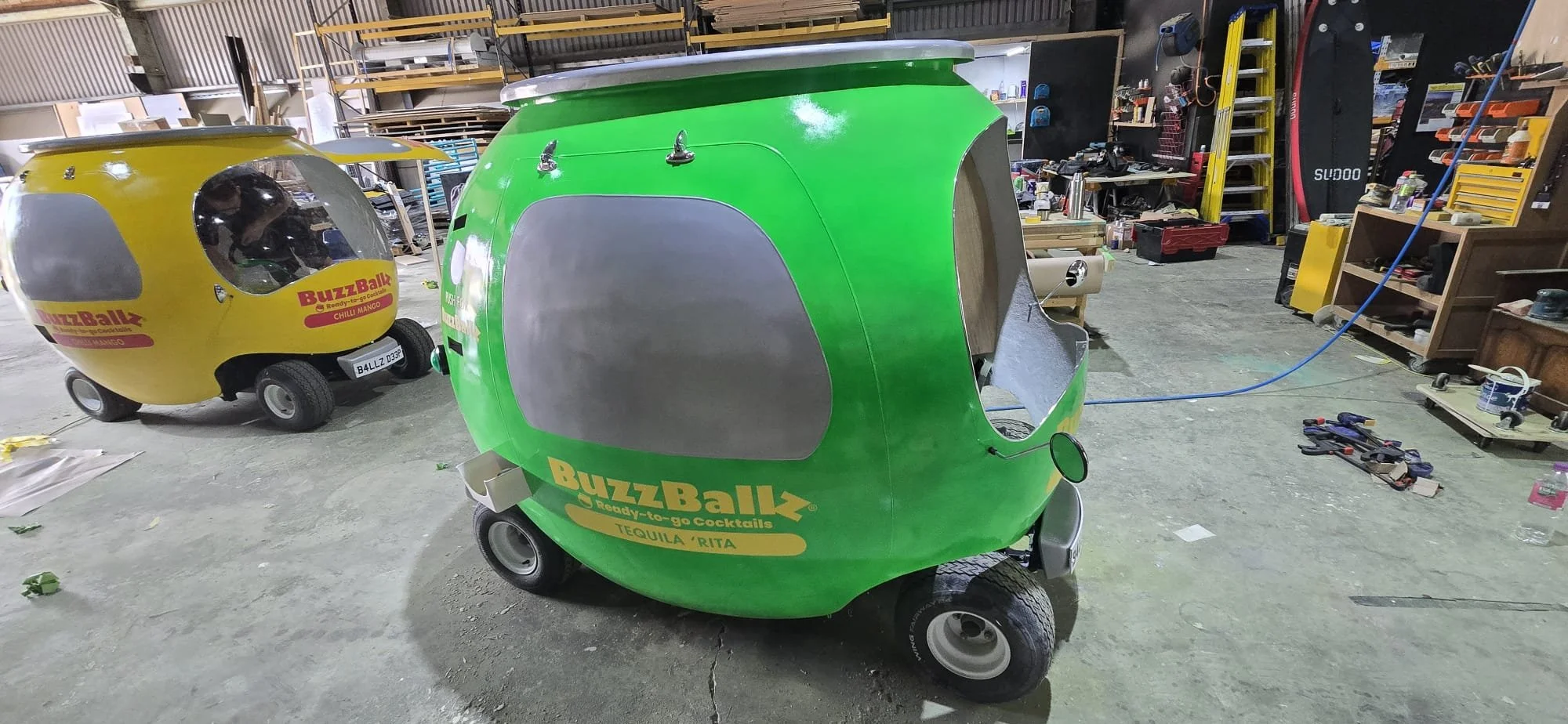 Two colorful, small, teardrop-shaped trailers or unicycle-like vehicles in a workshop. One yellow with a transparent bubble window, the other green with a closed side panel and a small wheel at front. There are tools and shelves around in the worksho