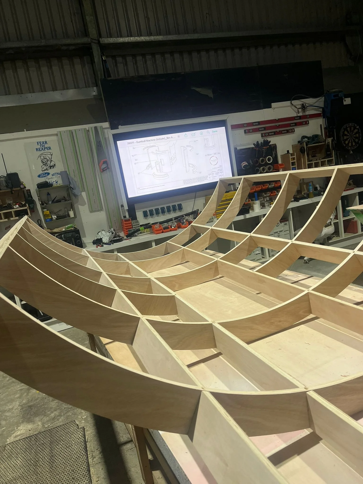 Wooden framework of THE BUZZBALLZ gatcha machine under construction in a workshop with tools and equipment in the background, a large screen displaying technical drawings.