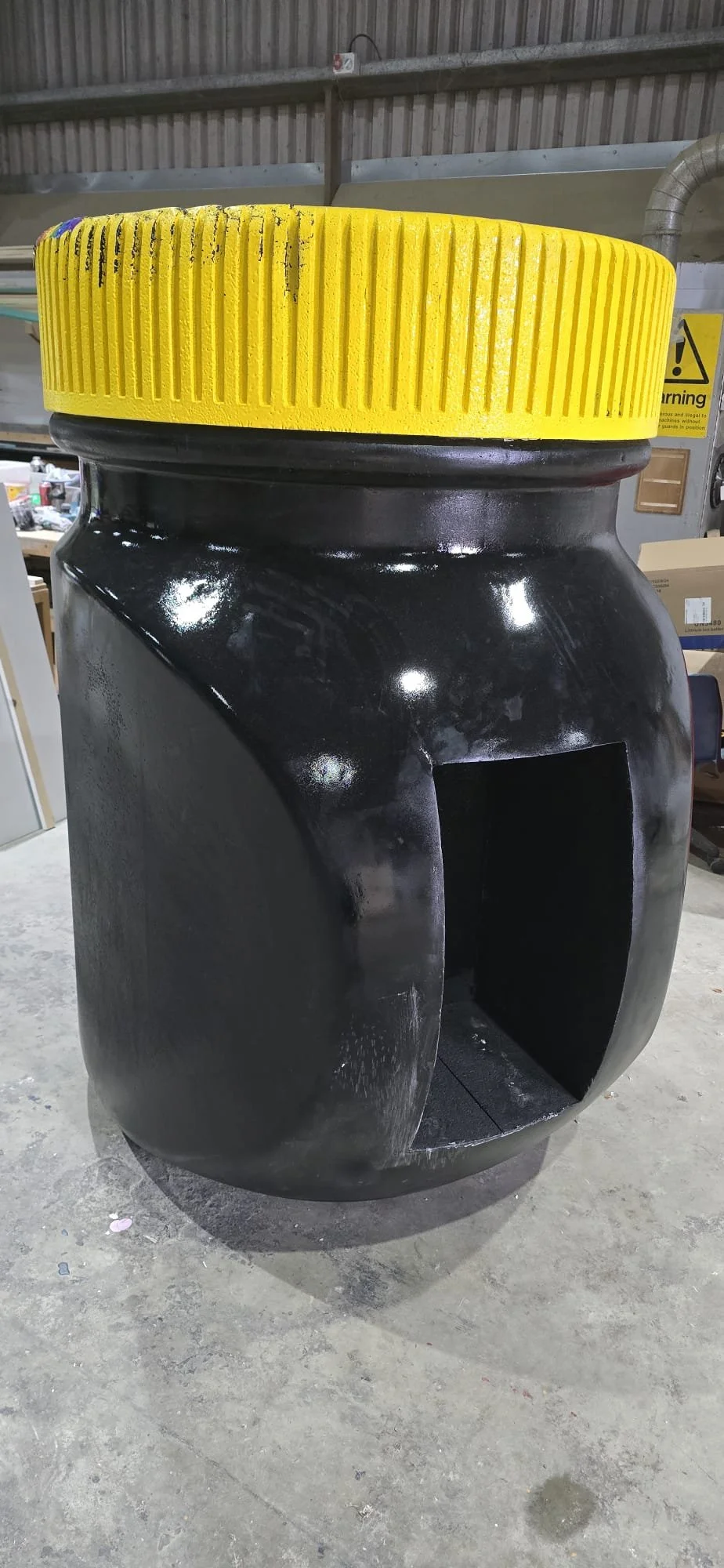 A black plastic trash bin with a large yellow lid, located on a concrete floor in a warehouse or storage area.