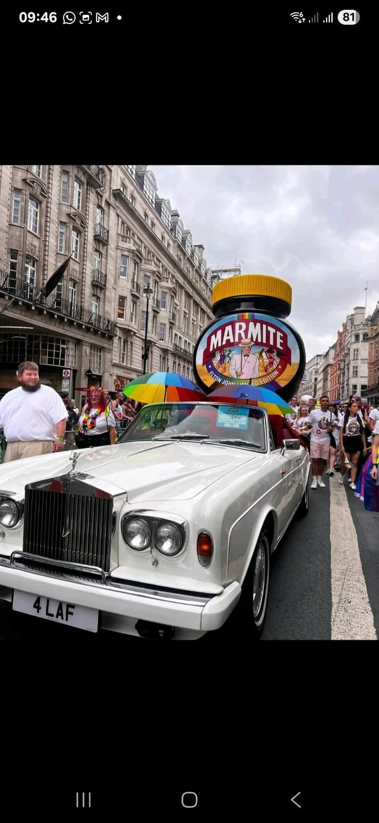 A vintage white car with a giant Marmite jar on the roof, surrounded by people holding umbrellas. The scene appears to be on a city street during a parade or festival.