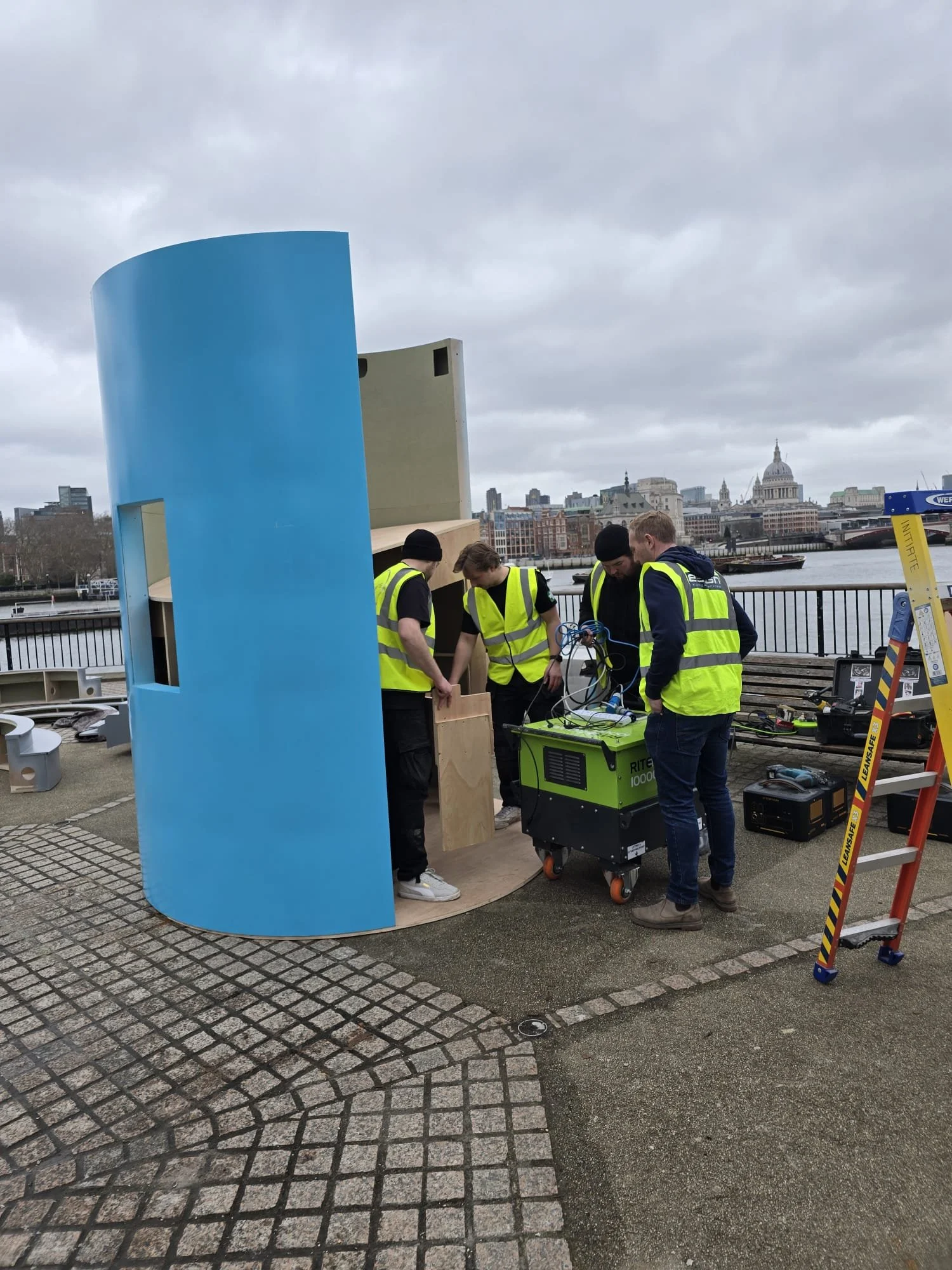 Four people in yellow safety vests working on equipment inside a curved blue structure near a river with city buildings in the background.
