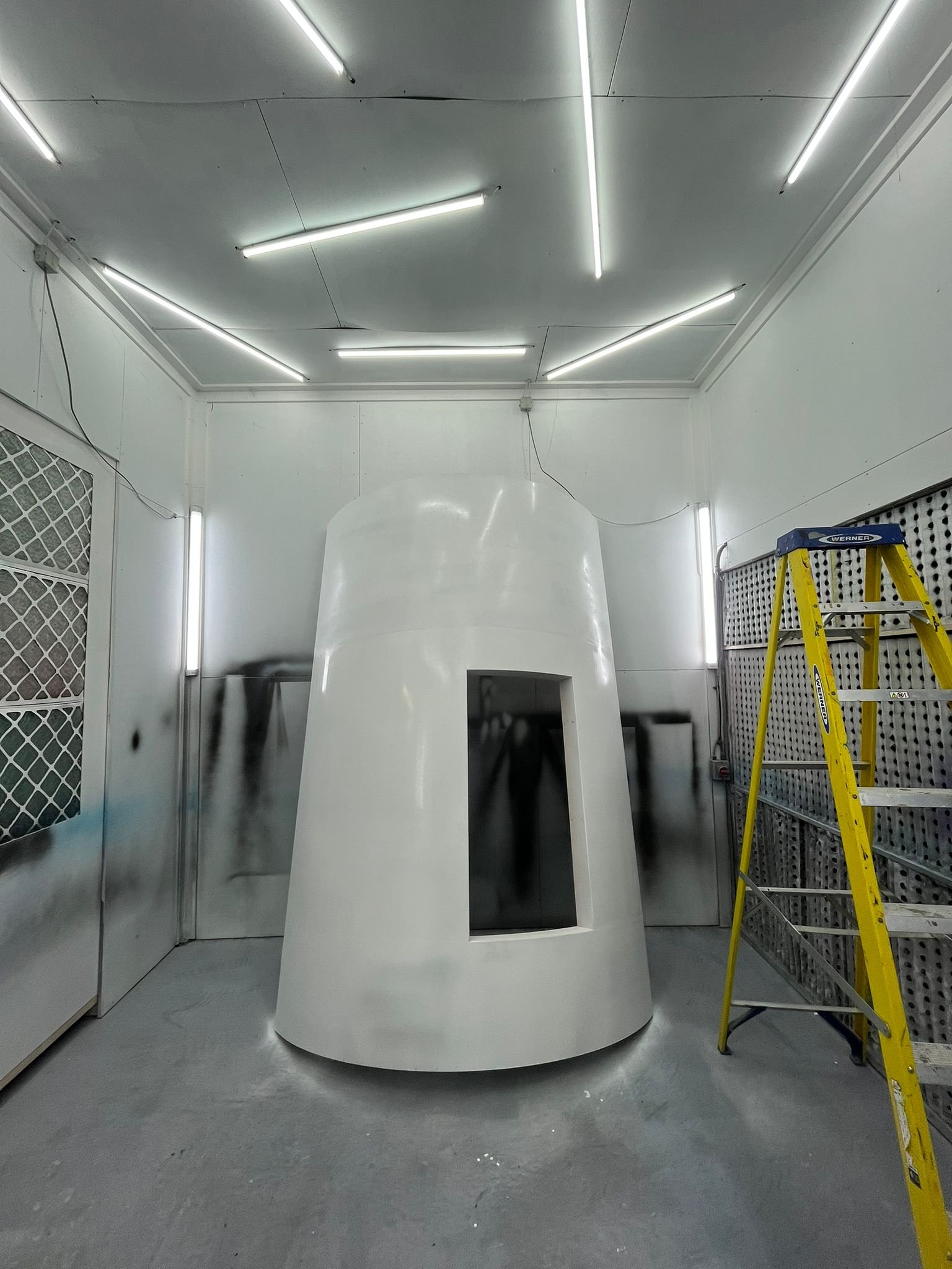 A white, spaceship-like structure with a rectangular window opening, inside a well-lit industrial or workshop space with metal walls and a yellow step ladder nearby.