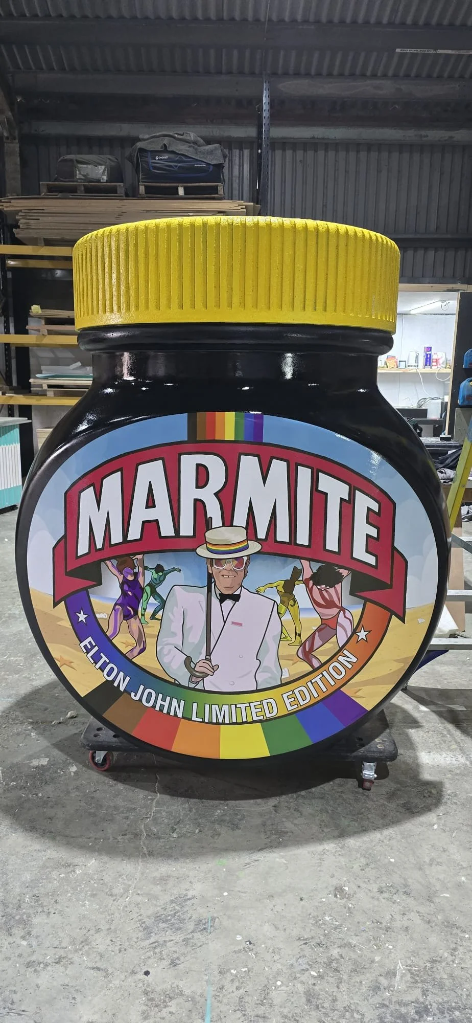 A large container with a Marmite label featuring Elton John as part of a limited edition. The label has a rainbow border and shows Elton John in a white suit and hat, surrounded by colorful dancing figures, set against a beach background.