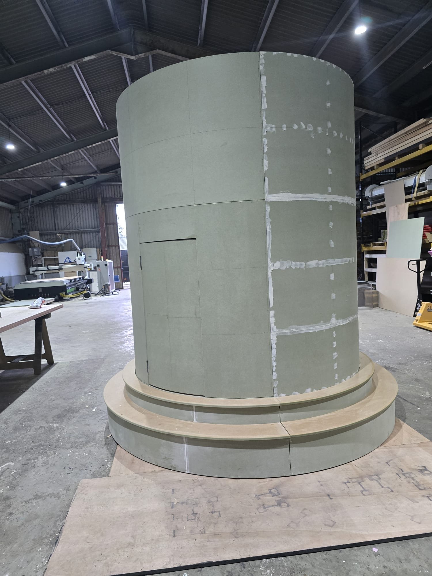 A large cylindrical structure under construction inside a woodworking workshop, situated on a circular platform with a layered wood base.