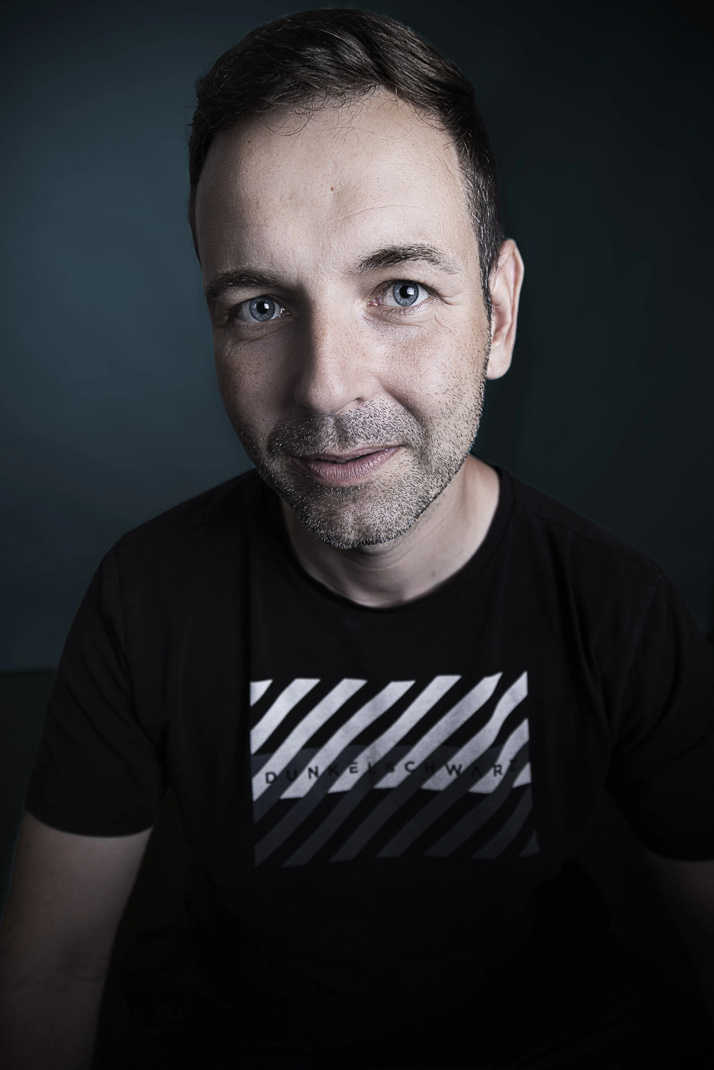 Porträt eines lachenden Mannes mit blauen Augen, dunklem Haar und Bart, trägert ein schwarzes T-Shirt mit weißen Streifen und Text, dunkler Hintergrund.