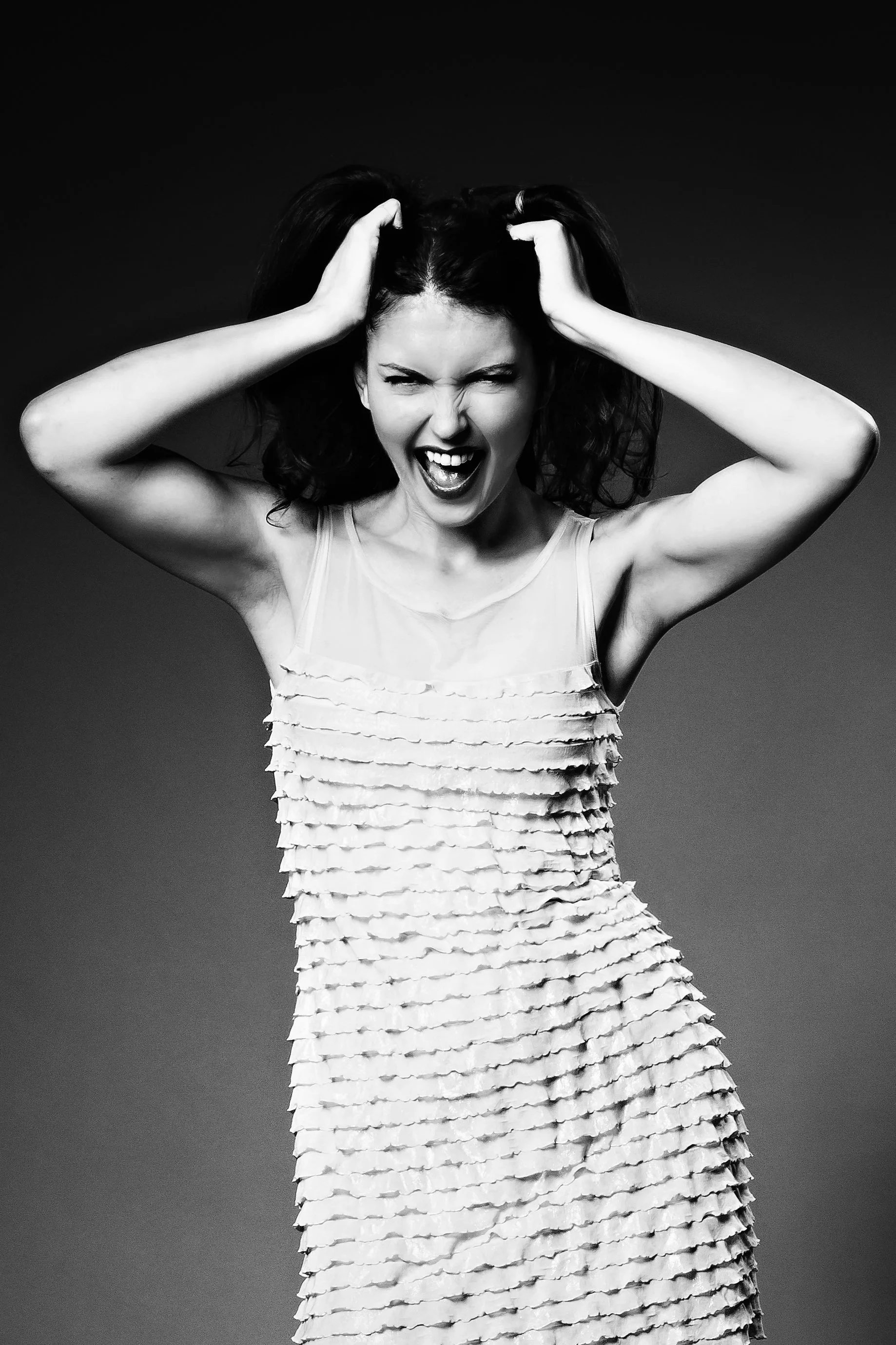 Schwarz-weiß Bild einer jungen Frau mit herausfordernder Haltung, die ihre Haare zerzaust und raubt. Sie trägt ein Kleid mit mehreren Schichten und hat einen energischen Gesichtsausdruck.