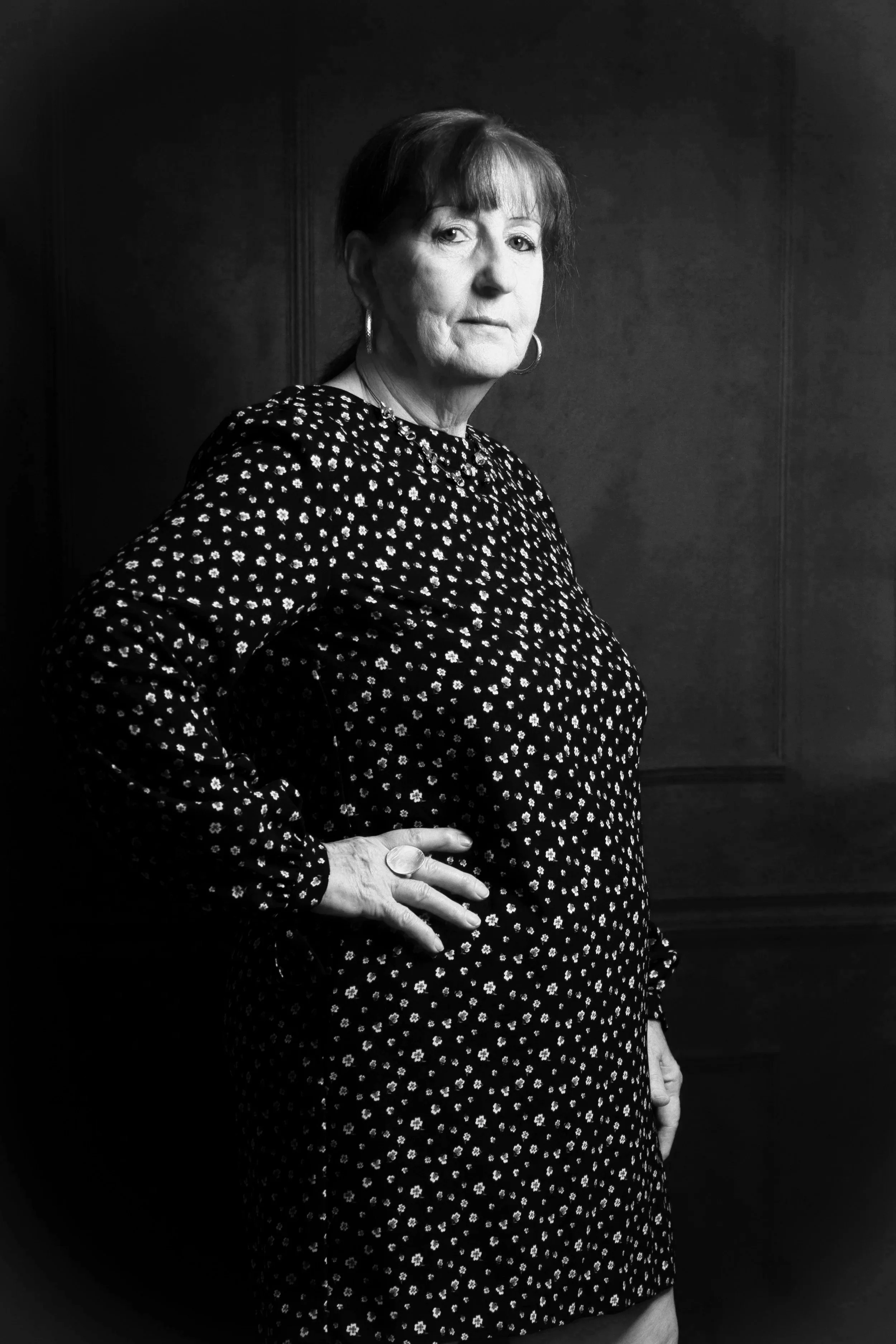 Schwarz-Weiß-Foto einer älteren Frau mit kurzen Haaren, die eine dunkle Bluse mit kleinen weißen Blumen trägt, vor einer dunklen Wand, mit ernstem Ausdruck und Hand auf der Hüfte.