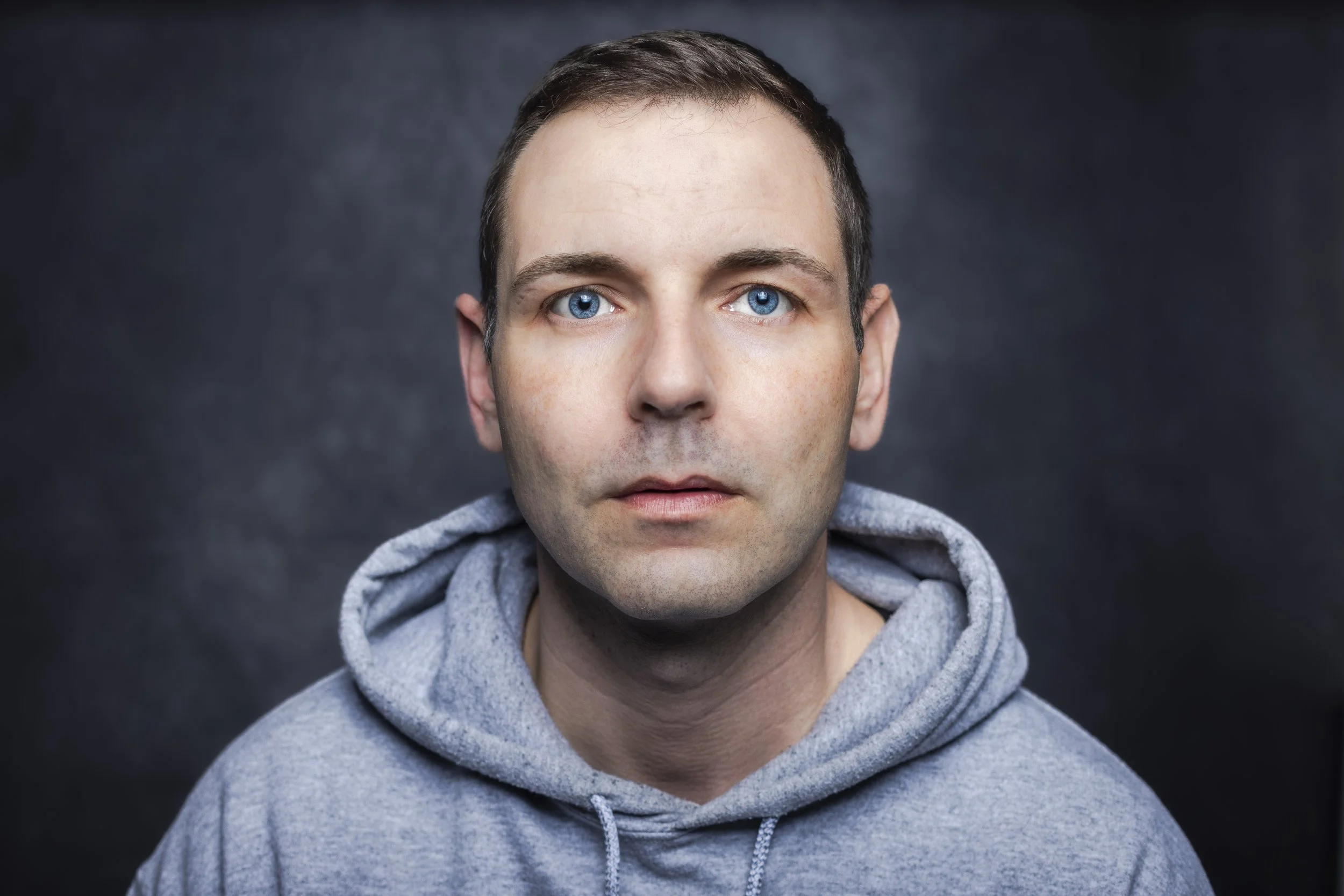 Nahaufnahme eines Mannes mit blauen Augen und dunklem kurzen Haar, trägt einen grauen Hoodie vor dunklem Hintergrund.