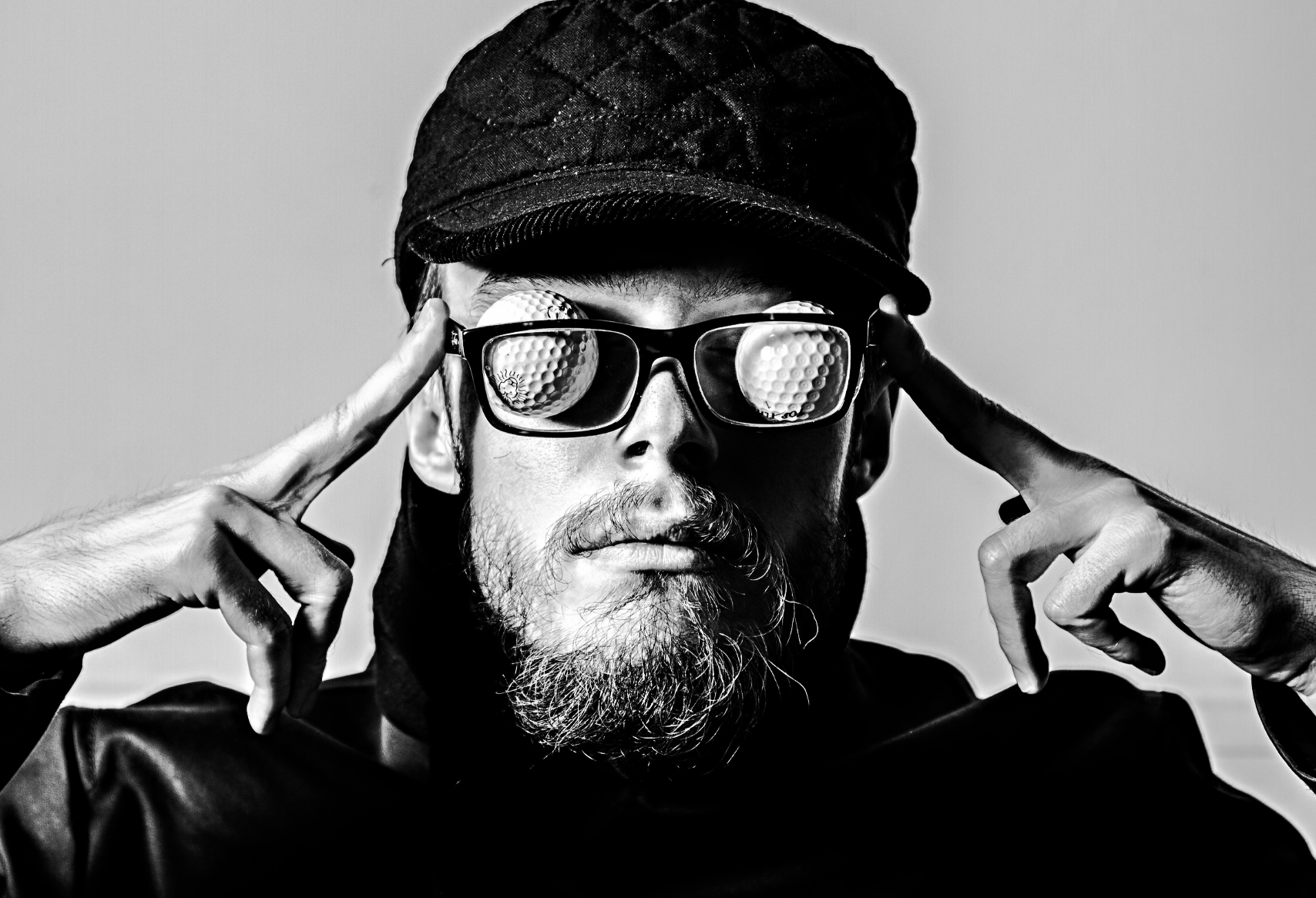 Schwarz-weiß-Porträt eines Mannes mit Bart, der eine Mütze und Sonnenbrille trägt, die reflektierende Gläser mit Golfball-Muster haben. Er berührt die Brille mit den Fingern.