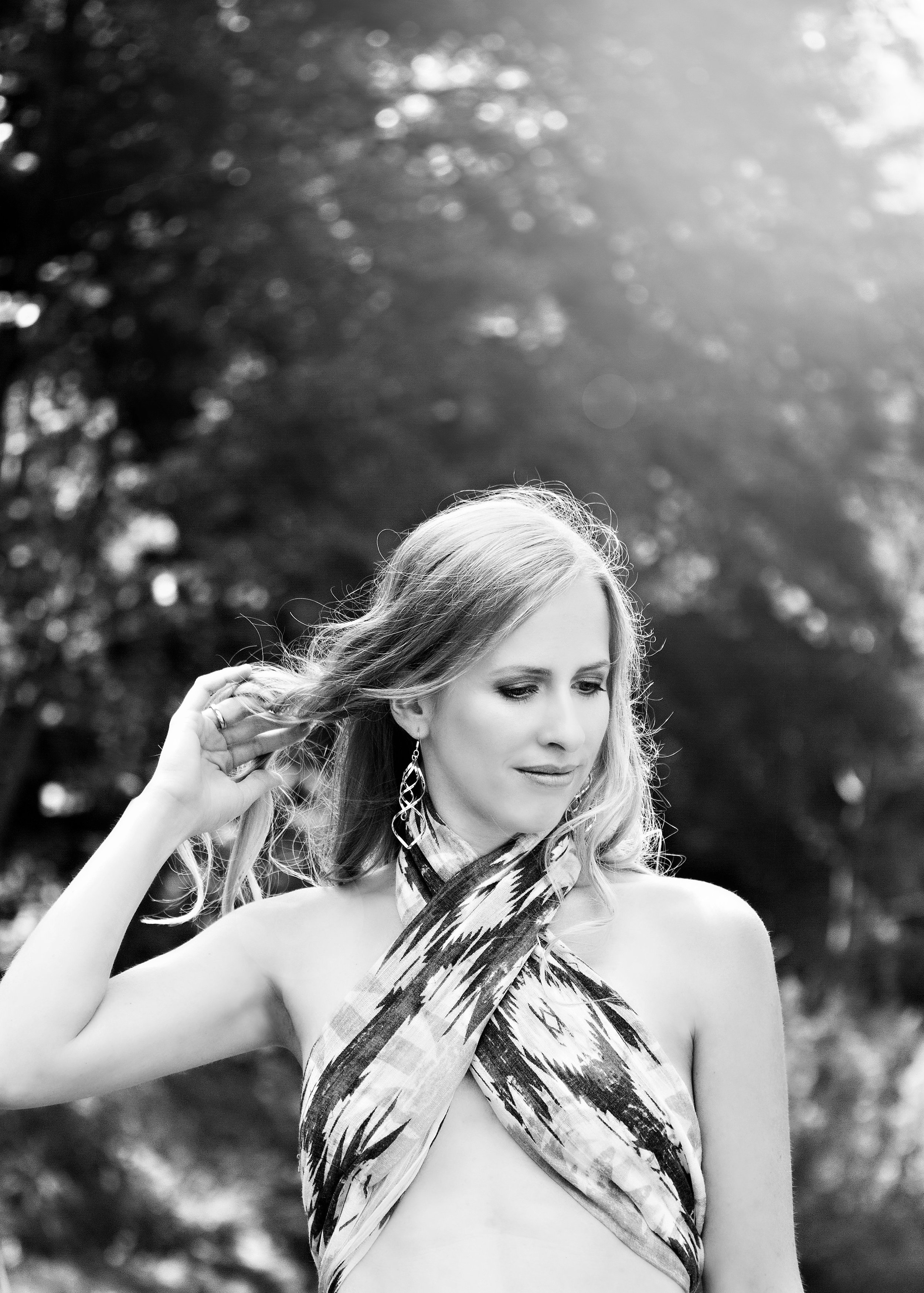 Schwarz-weiße Aufnahme einer Frau mit langem Haar, die an einer nassen Strickjacke oder einem Schal mit Wüstenmuster trägt, im Freien mit Bäumen im Hintergrund, bei Sonnenlicht.