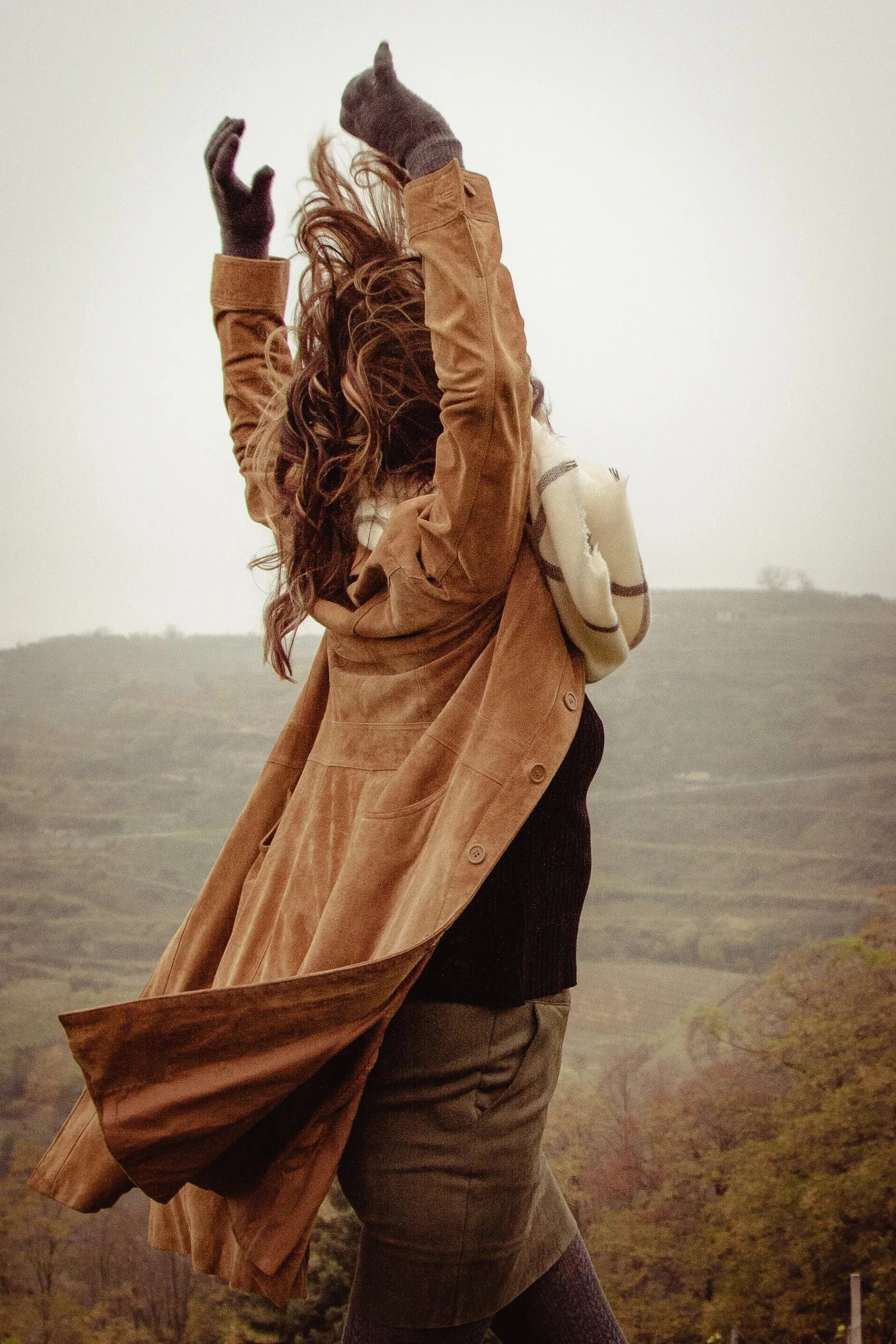 Frau mit lockigem Haar, die einen langen braunen Mantel trägt, hebt die Arme in der Luft, im Hintergrund eine hügelige Landschaft mit Bäumen.