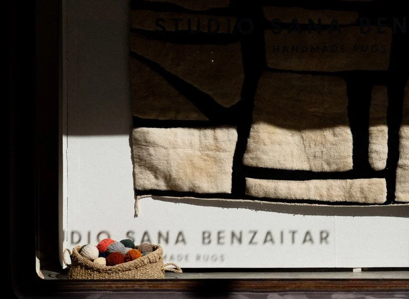Een tapijt met de tekst 'STUDIO SANA BENZAITAR HANDMADE RUGS' en een tas gevuld met verschillende gekleurde viltballen.